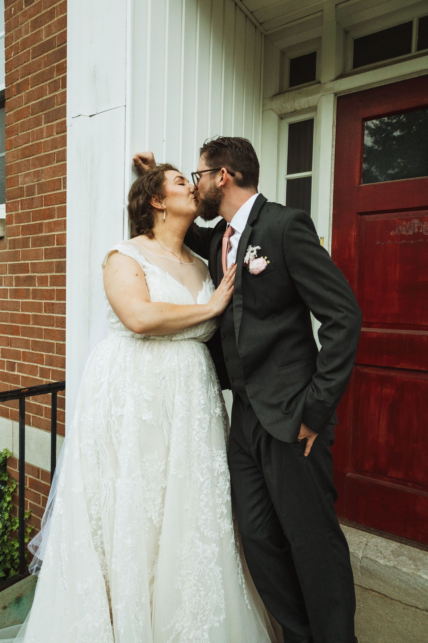 Romantic wedding couple sharing intimate kisses against a red brick wall with white trim.