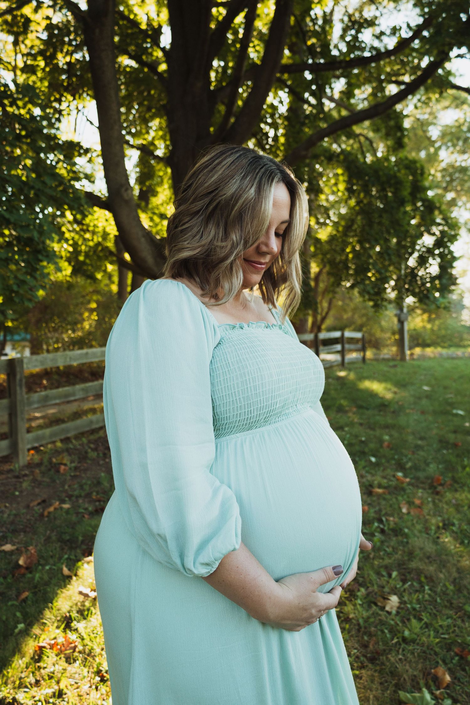 A person in a mint green dress cradles their belly while standing in a sunlit park with autumn leaves on the ground.
