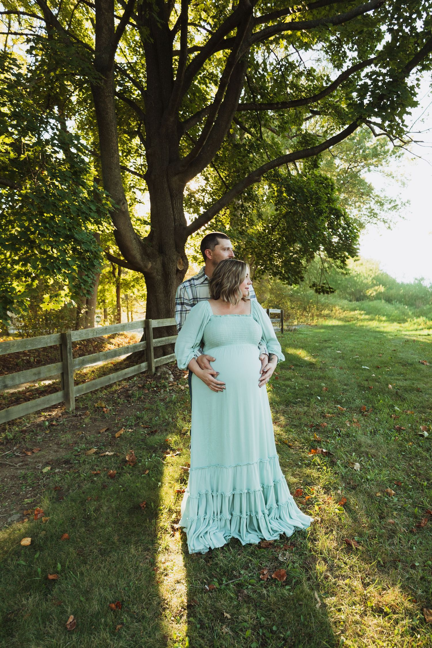 A person in a mint green maxi dress twirls in a sunlit garden setting with wooden fencing and trees in the background.