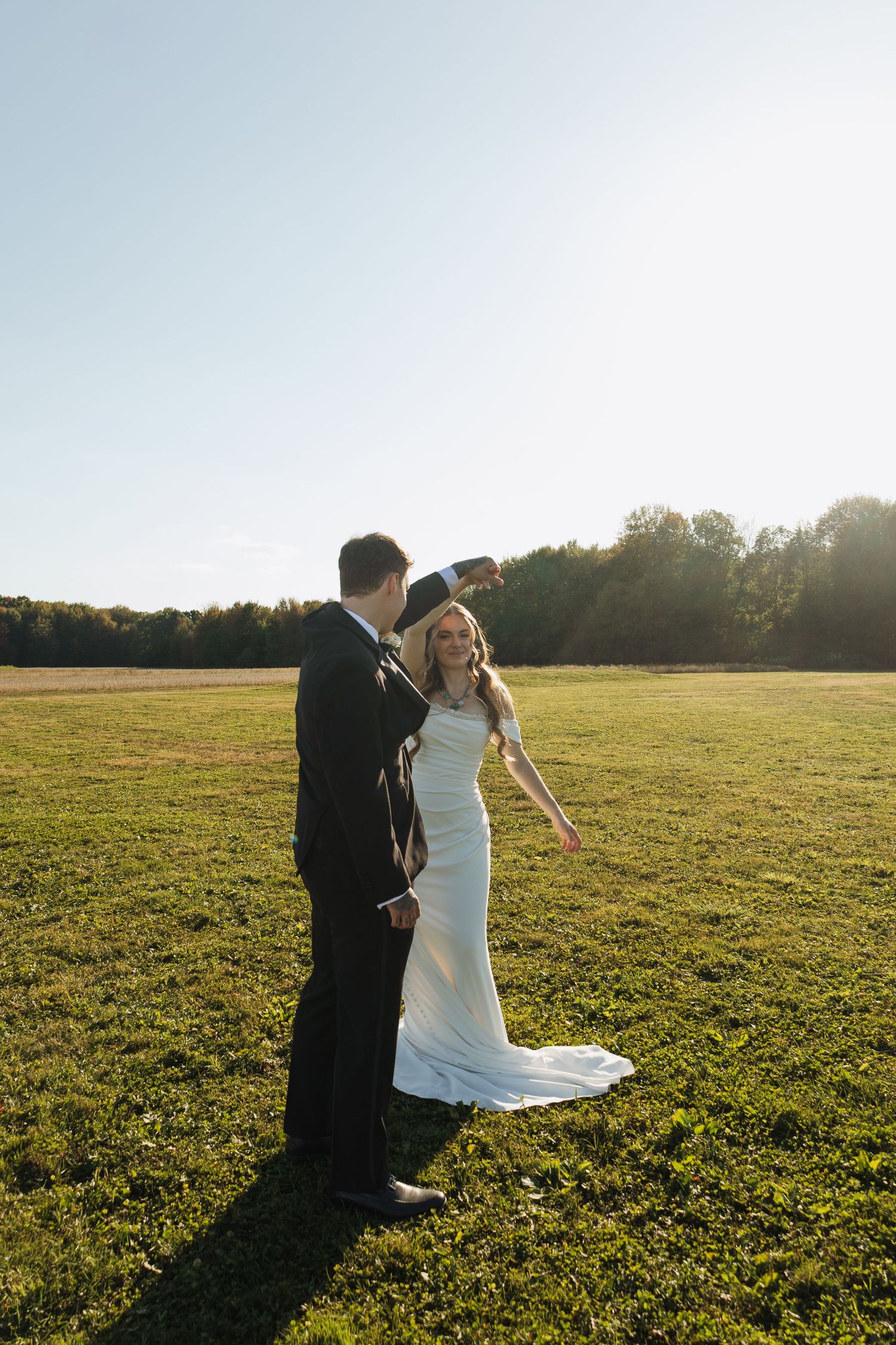 A couple in wedding attire posing in a sunlit open field during golden hour.