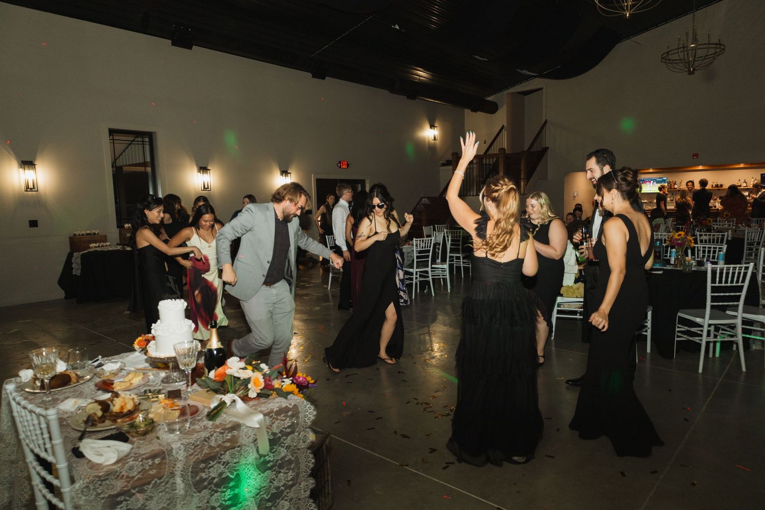 People dancing and celebrating at a wedding reception in a dimly lit venue with decorative lighting.