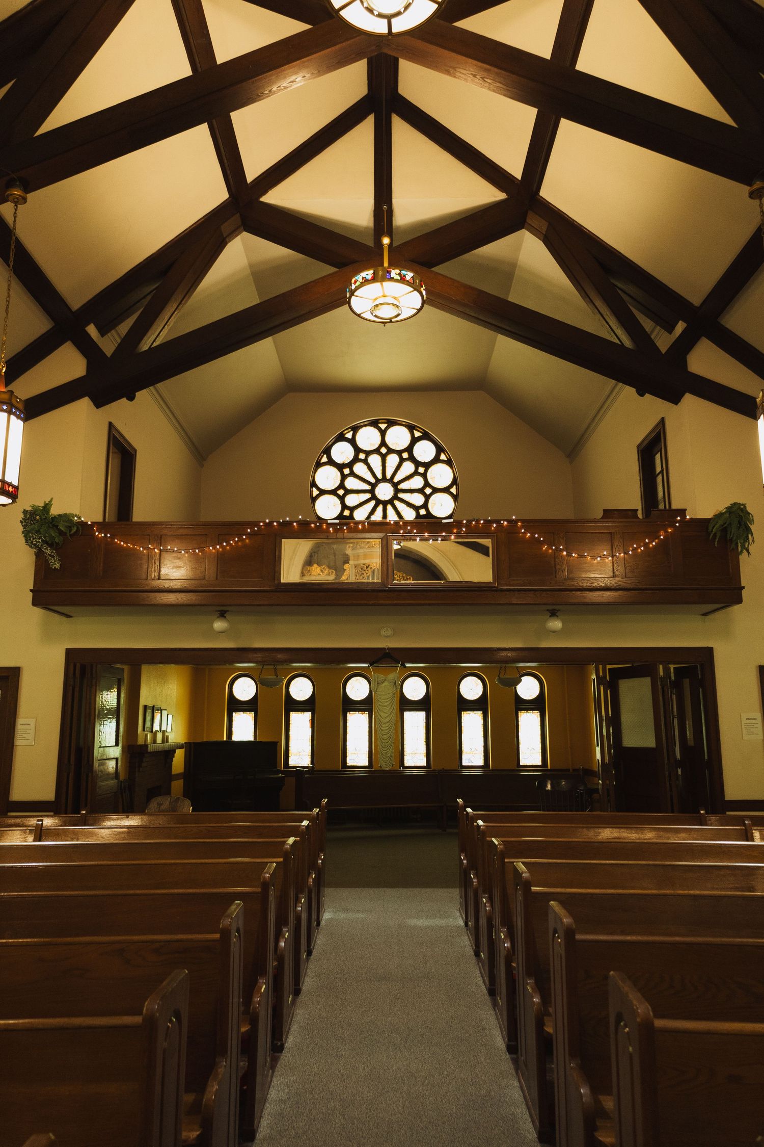 Elegant church interior with wooden pews, vaulted ceiling beams, rose window, and balcony decorated with holiday garland.
