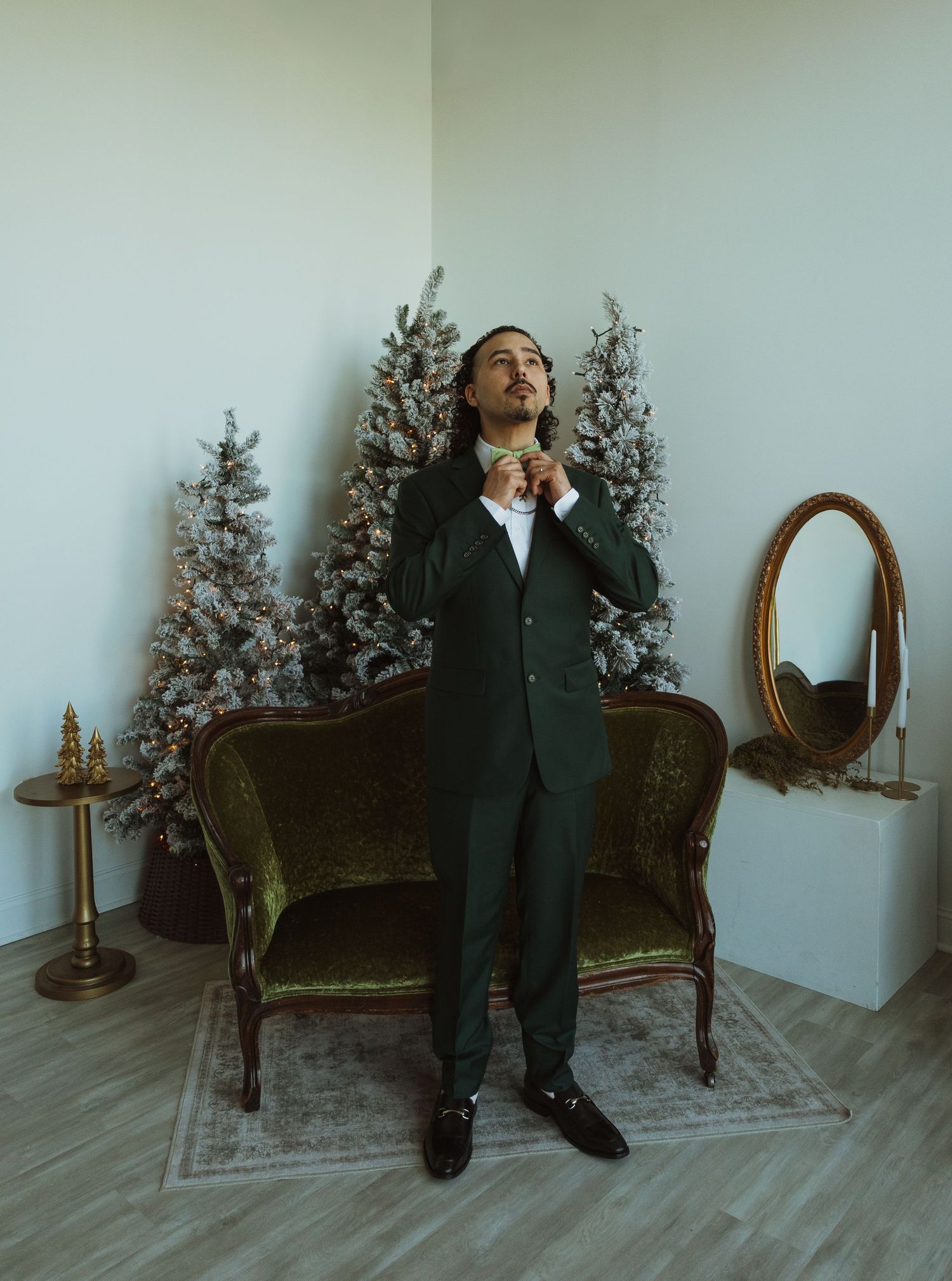 Groom in dark green suit adjusts bow tie beside flocked Christmas trees and vintage velvet sofa.