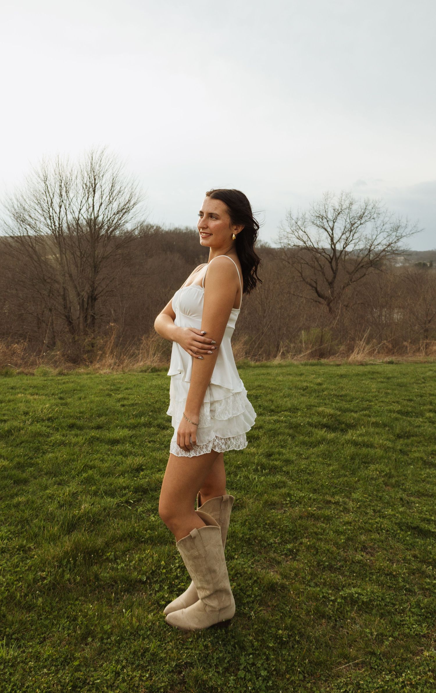 Young woman in white lace romper and cowboy boots standing on green grass field with bare trees in background.