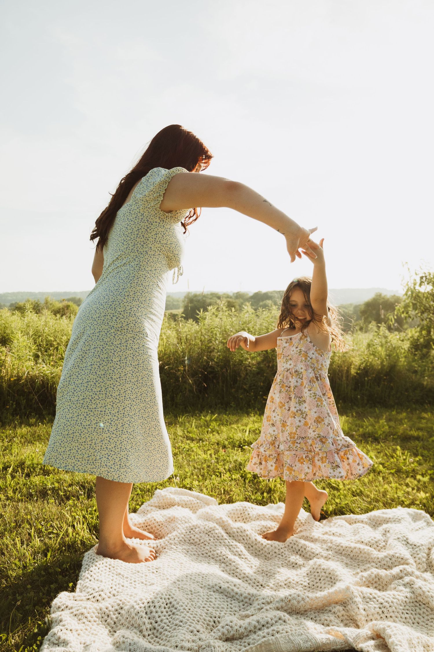 A parent and child dance together on a white blanket in a sunny field at sunset.