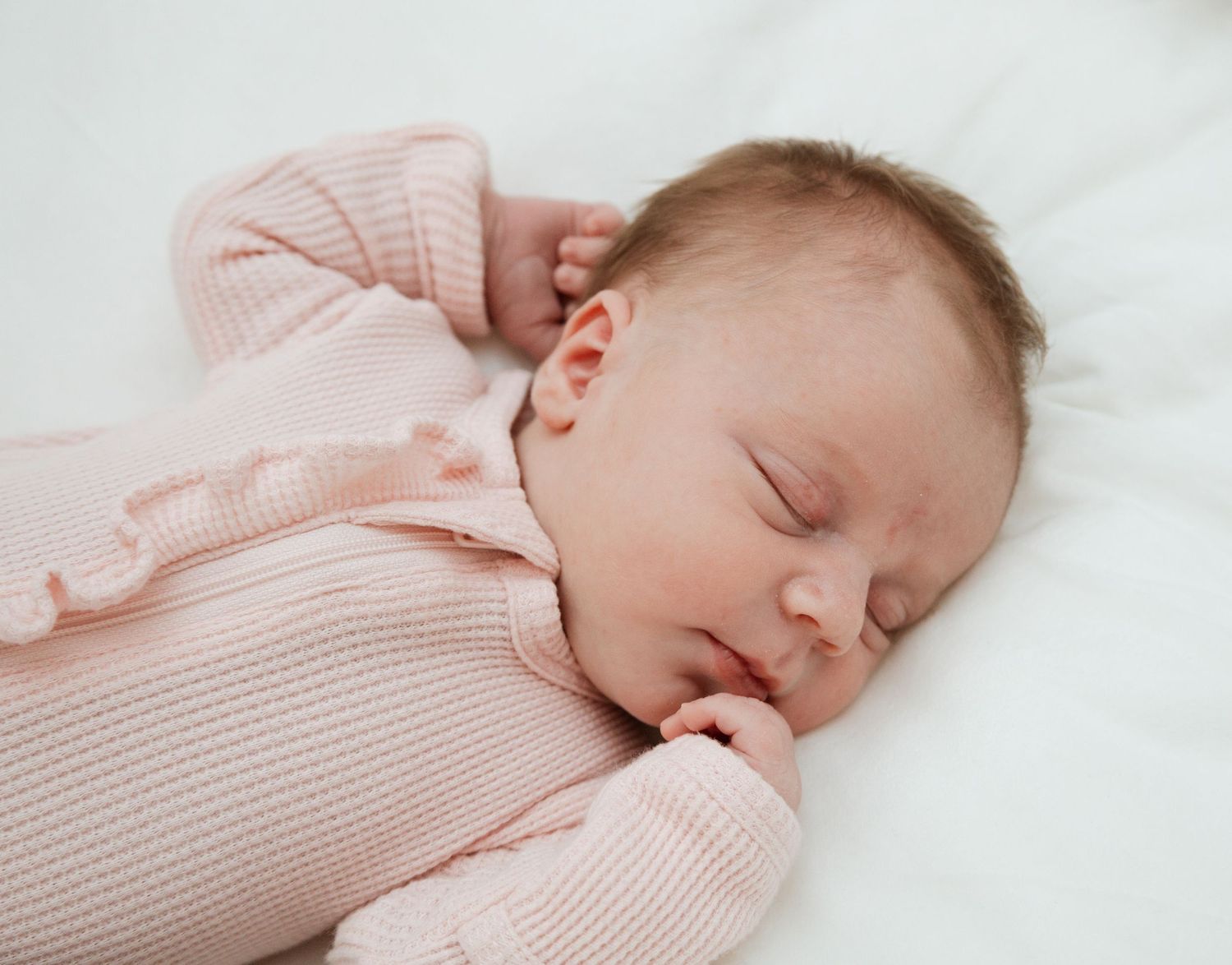 A peaceful sleeping baby wearing a soft pink knit sweater rests against white bedding.
