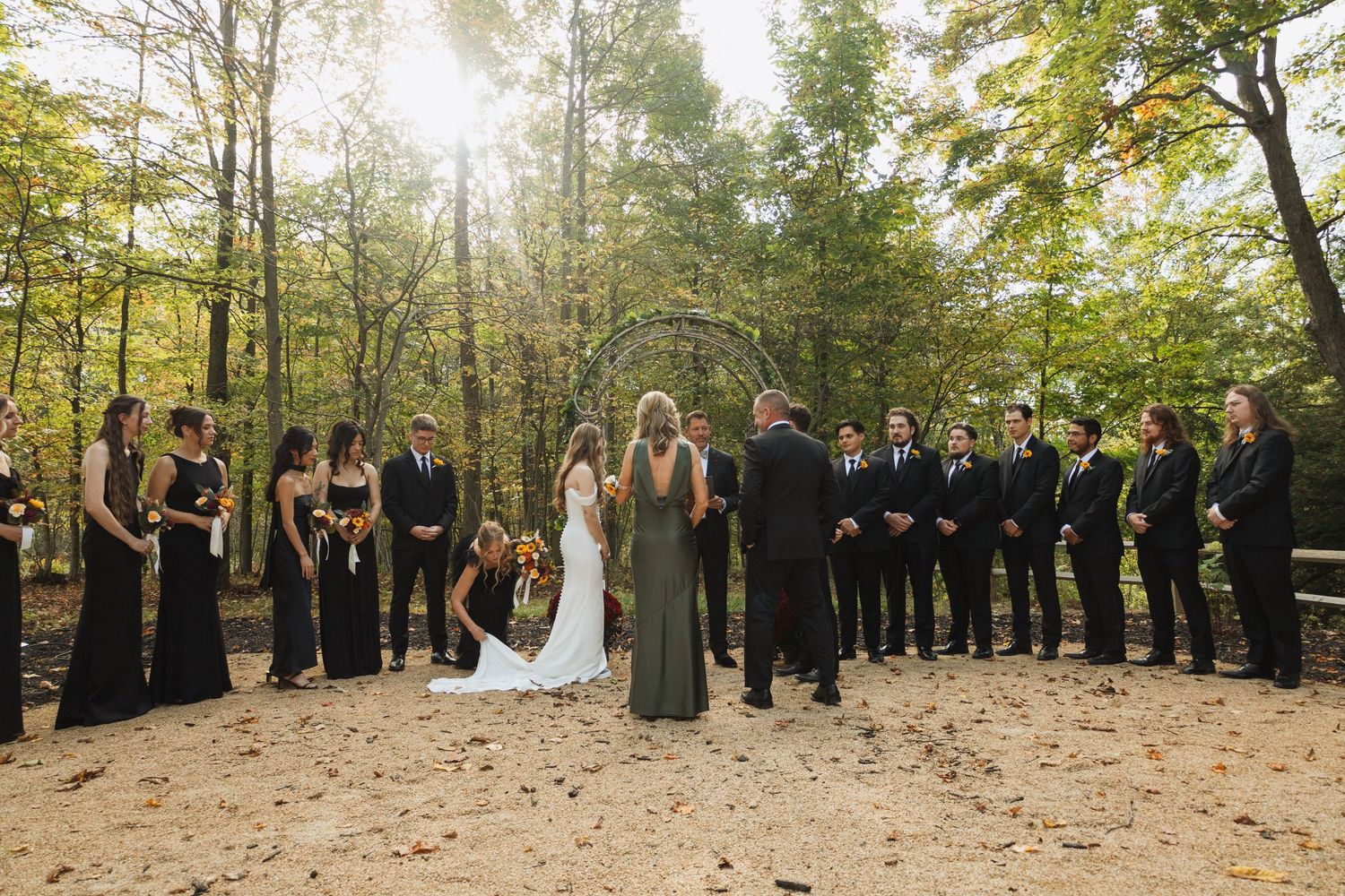 Outdoor wedding ceremony in a sunlit forest with wedding party in formal attire.
