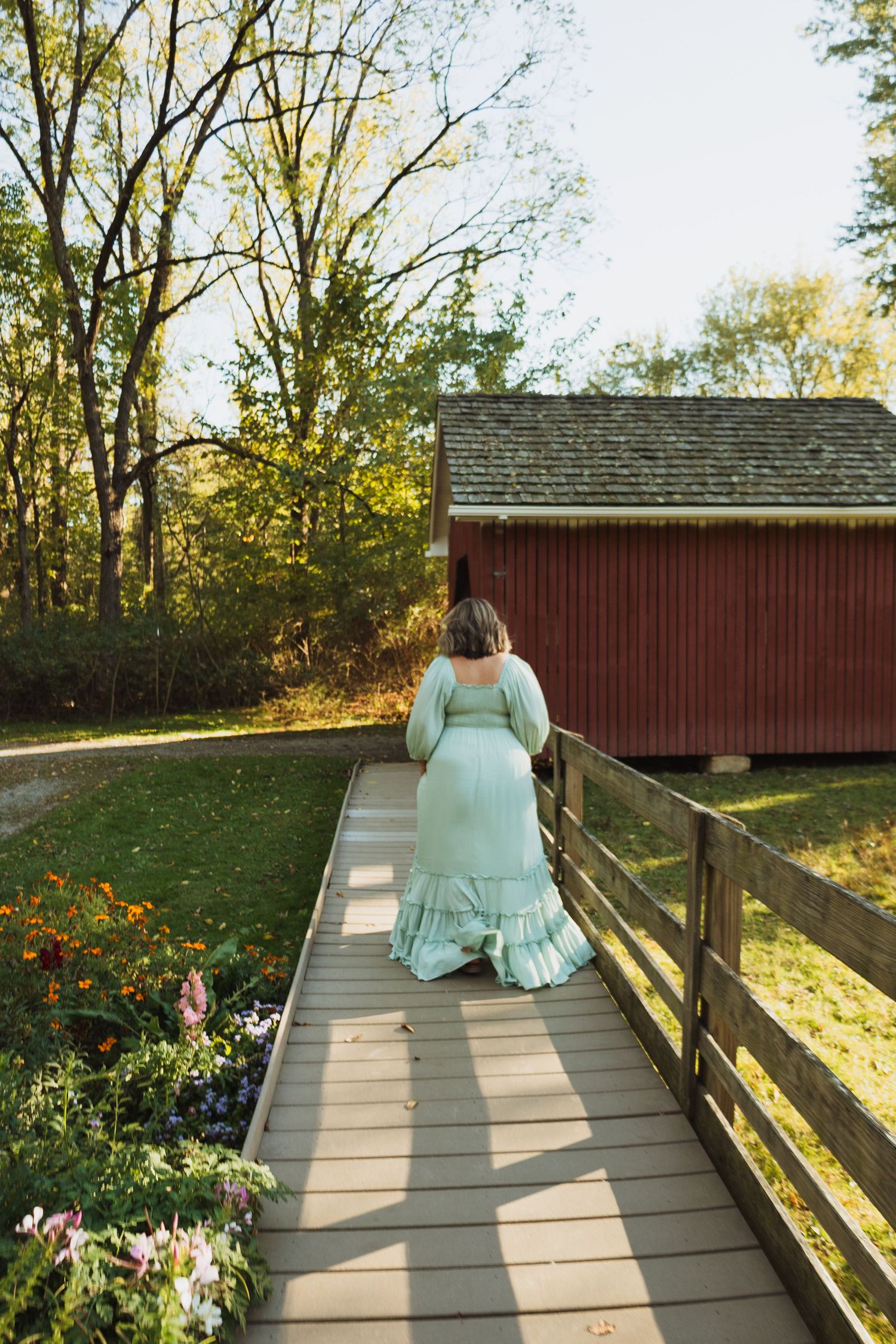 A person in a mint green dress sits peacefully on a wooden boardwalk surrounded by autumn foliage and flowers.