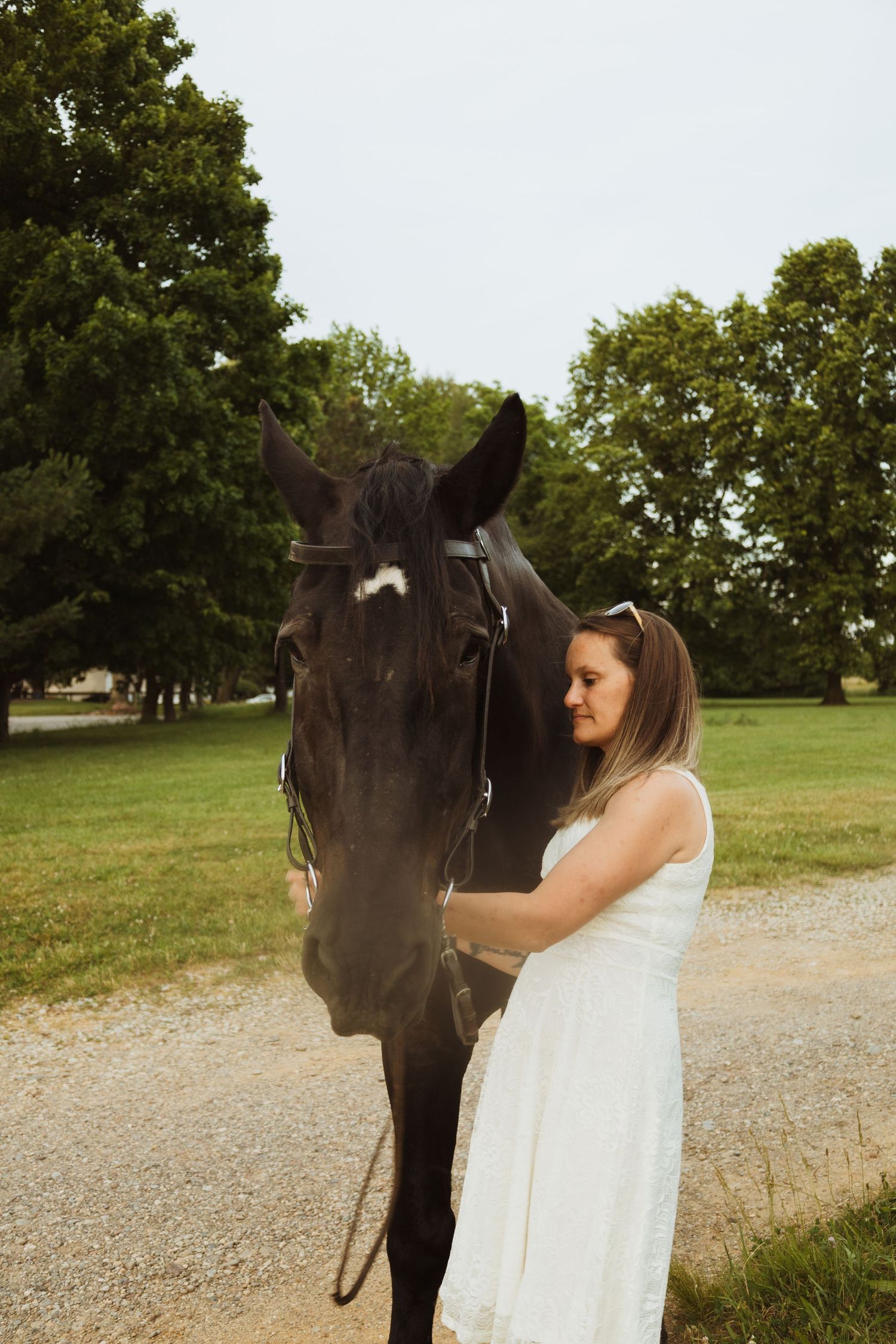 A woman in a white dress tenderly stands beside a dark horse in a lush green park setting.