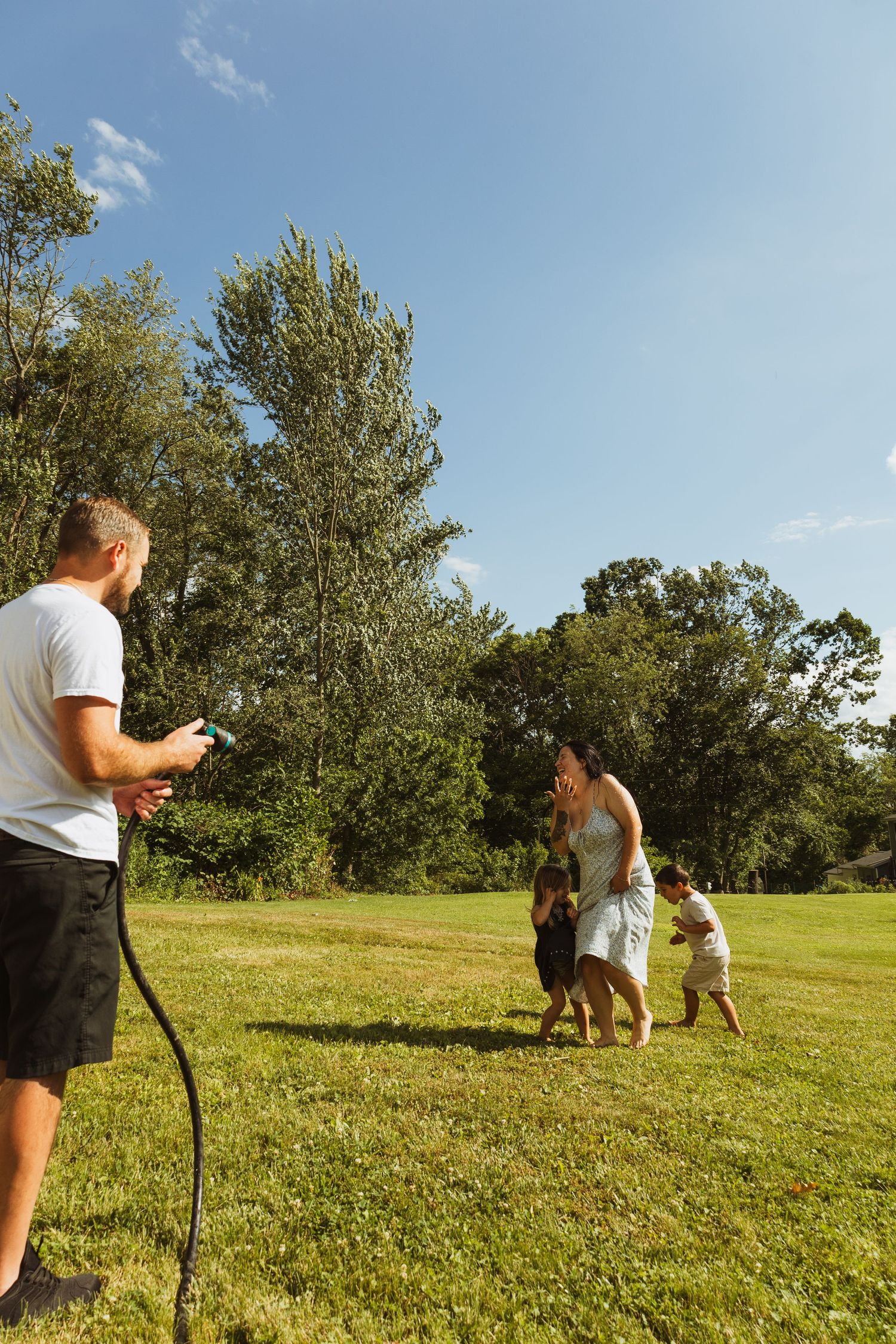 Family playing with garden hose on sunny day in green backyard, man spraying water at woman and children near trees.