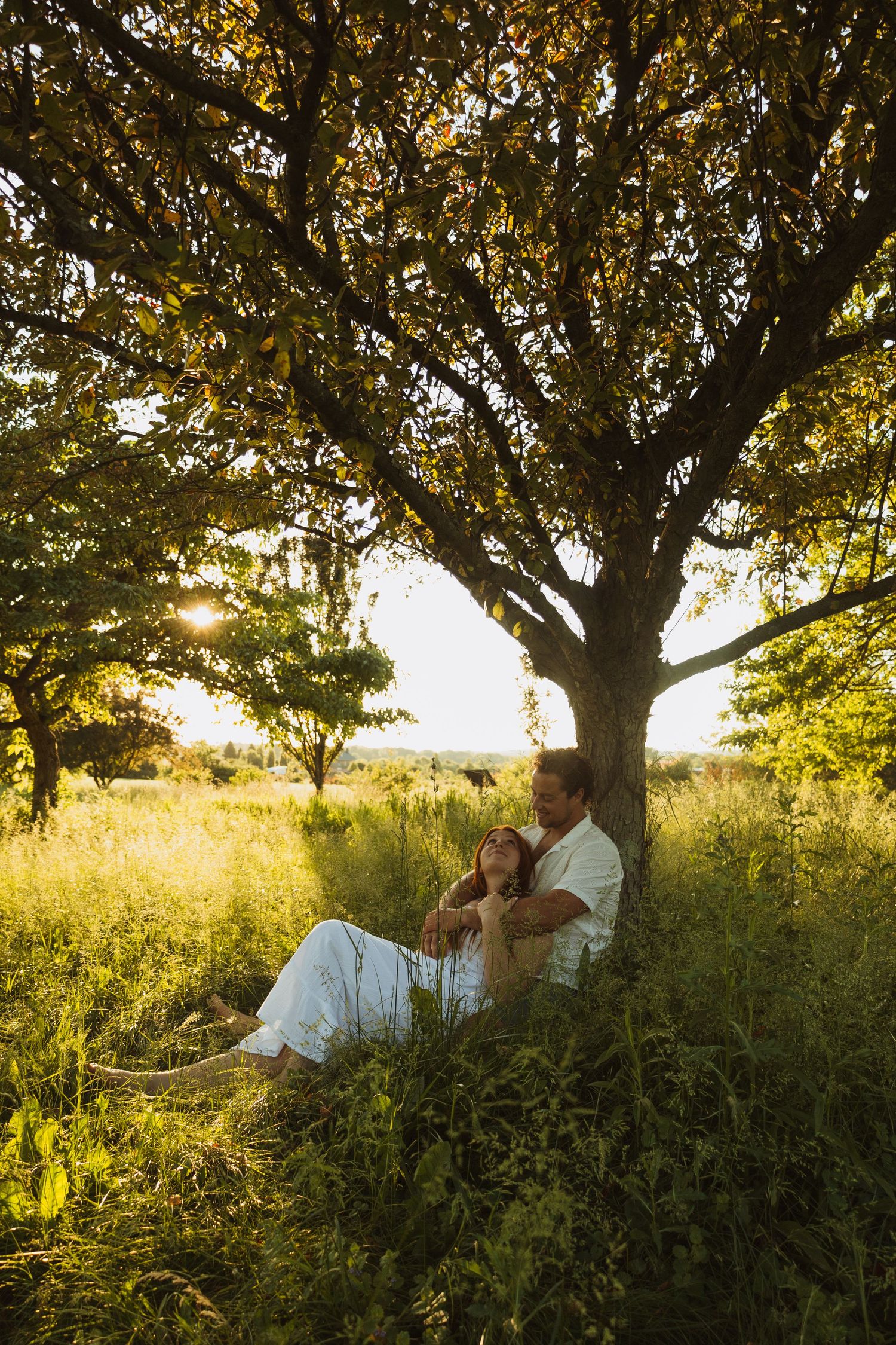 Couple in white sitting under a tree in a sunlit green meadow, enjoying a peaceful moment together.