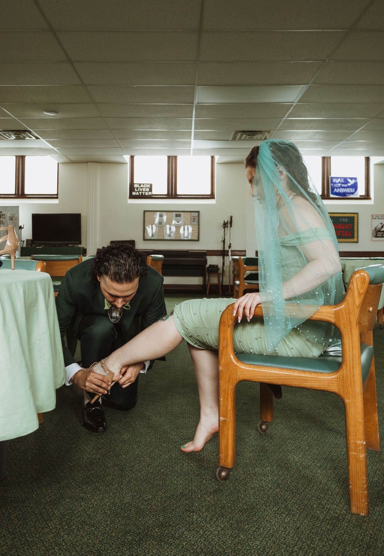 Groom kneeling to help bride with shoes as she sits in chair wearing green dress and teal veil.