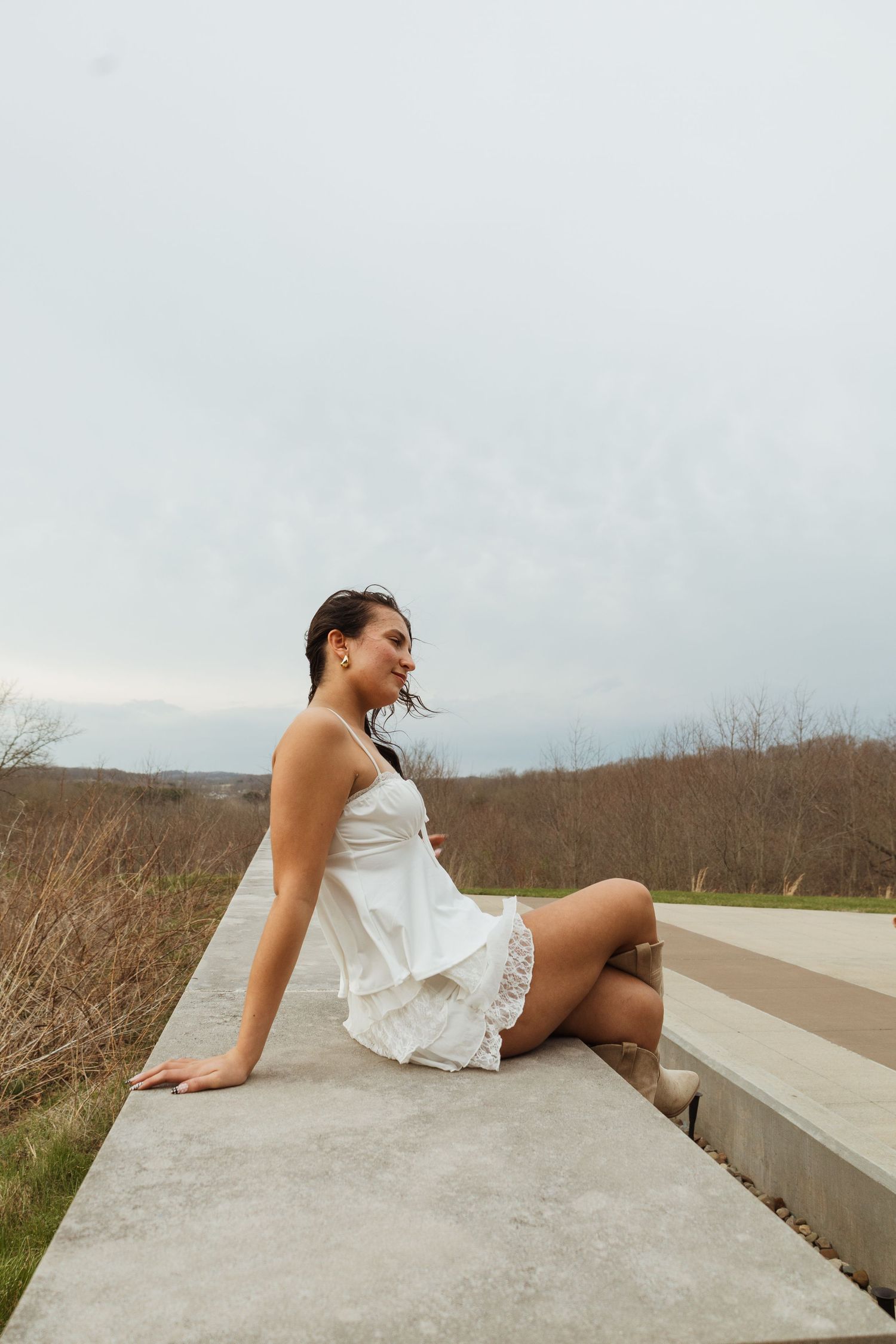A young woman in a white mini dress sits gracefully on a concrete ledge outdoors with a cloudy sky backdrop.
