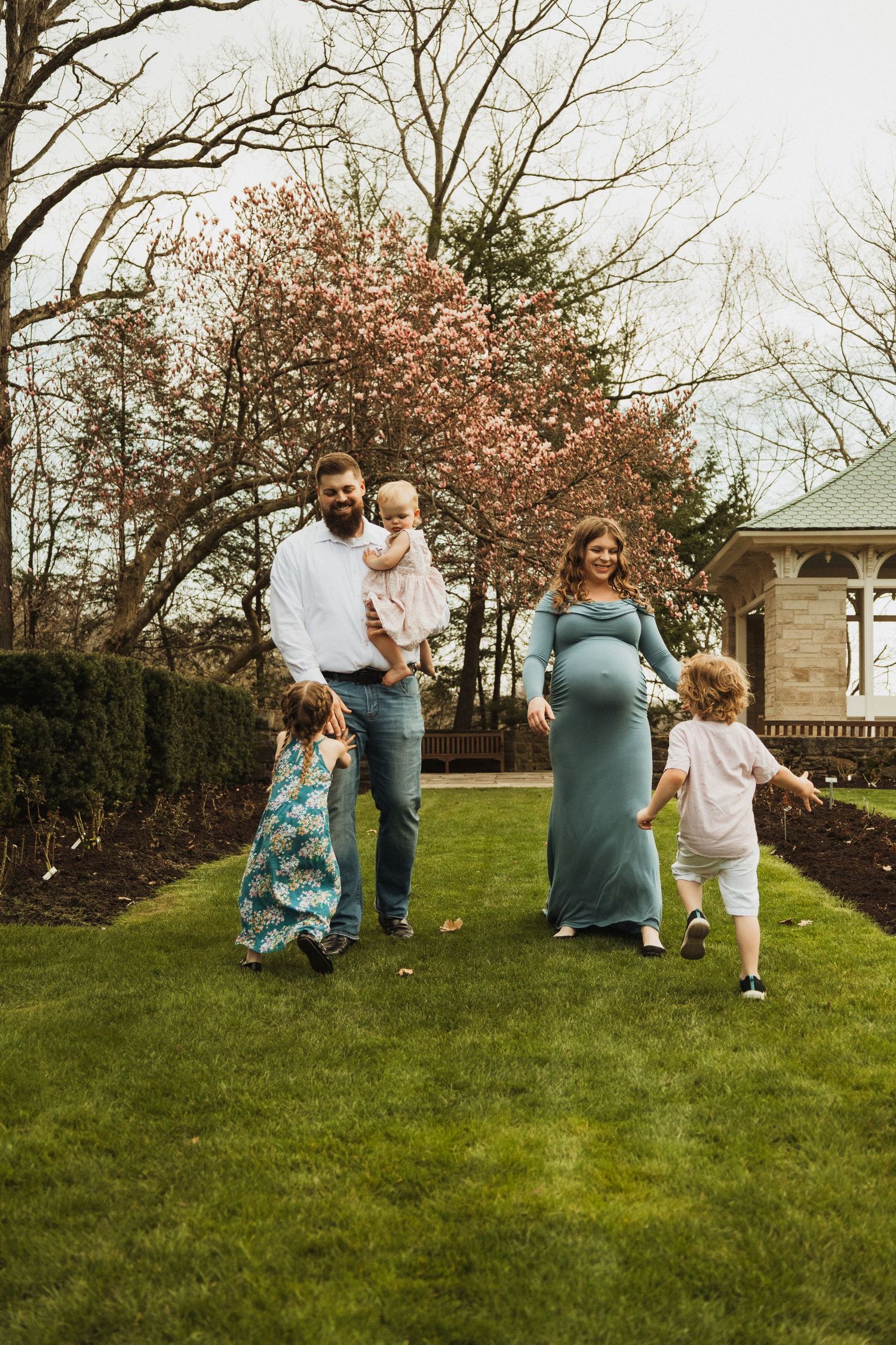 Family walking together on a spring lawn with blooming trees and a gazebo in the background.