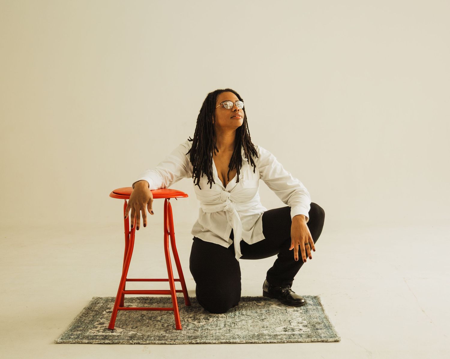 Creative studio pose with person crouching next to red stool on patterned area rug.