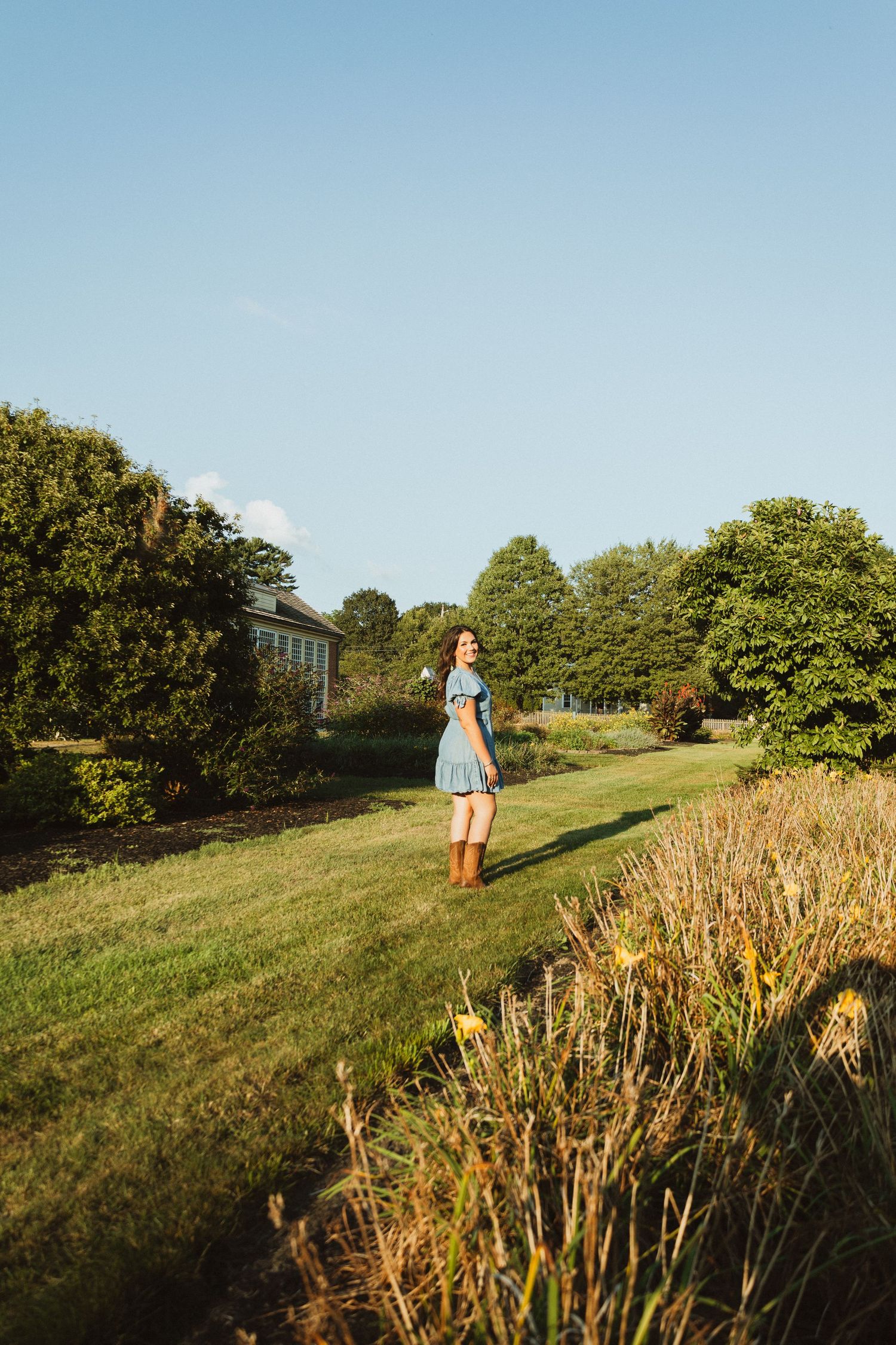 A person stands on a grassy hillside path at golden hour with trees and vegetation surrounding them in summer.