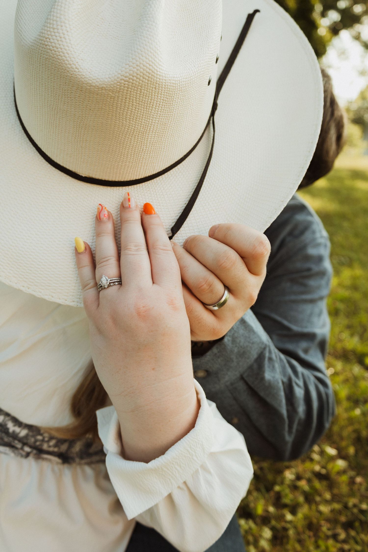 Close-up of hands wearing engagement ring and holding white cowboy hat against natural backdrop.