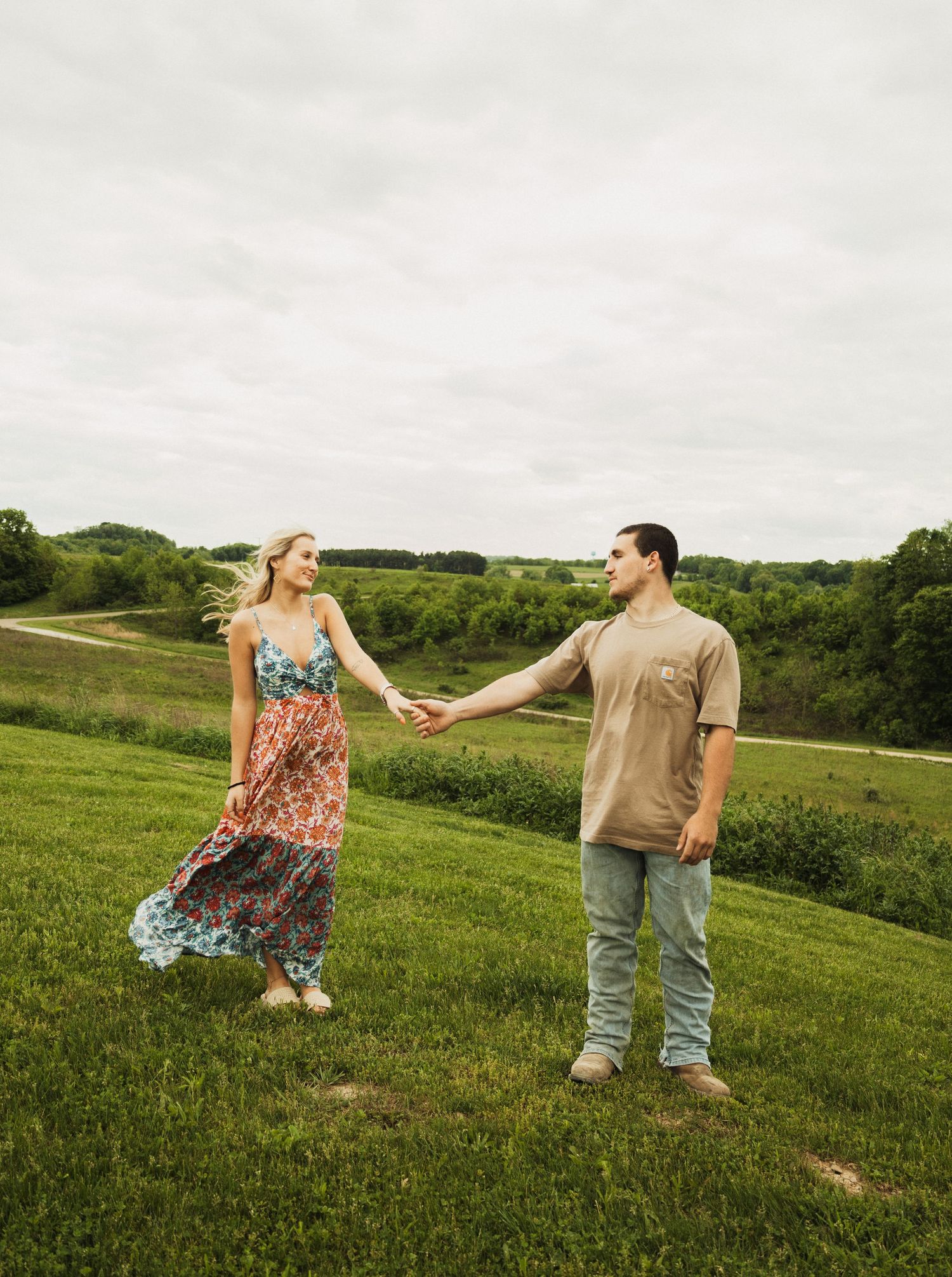 A couple in casual attire holding hands while standing apart on a green hillside with rolling terrain in the background.