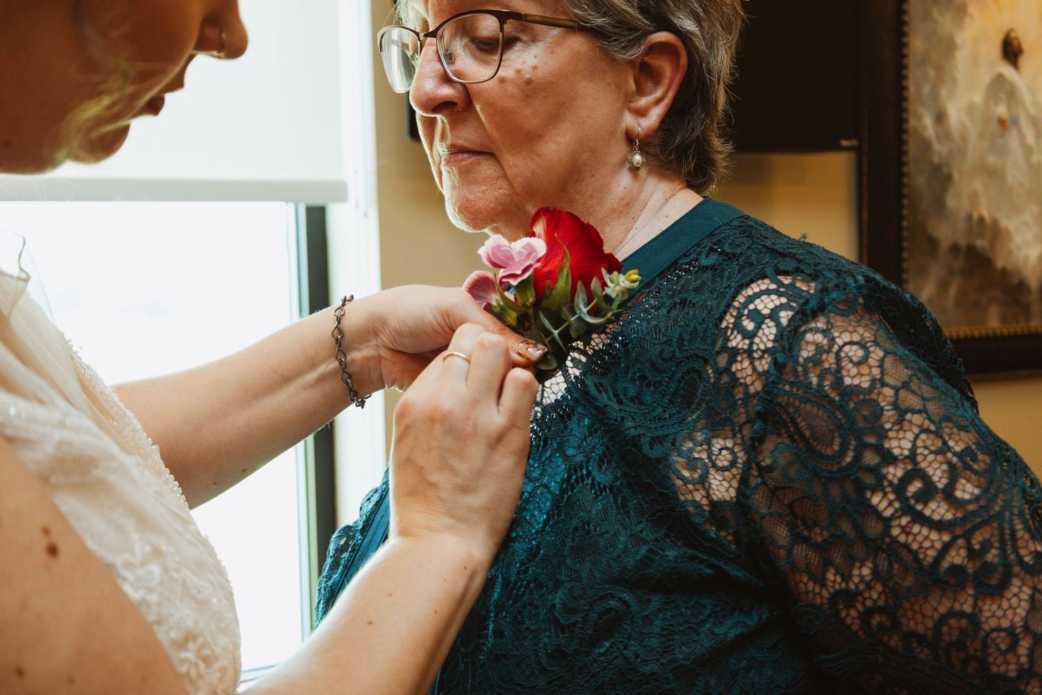Someone pins a red rose boutonniere onto a person wearing an elegant dark teal lace dress.