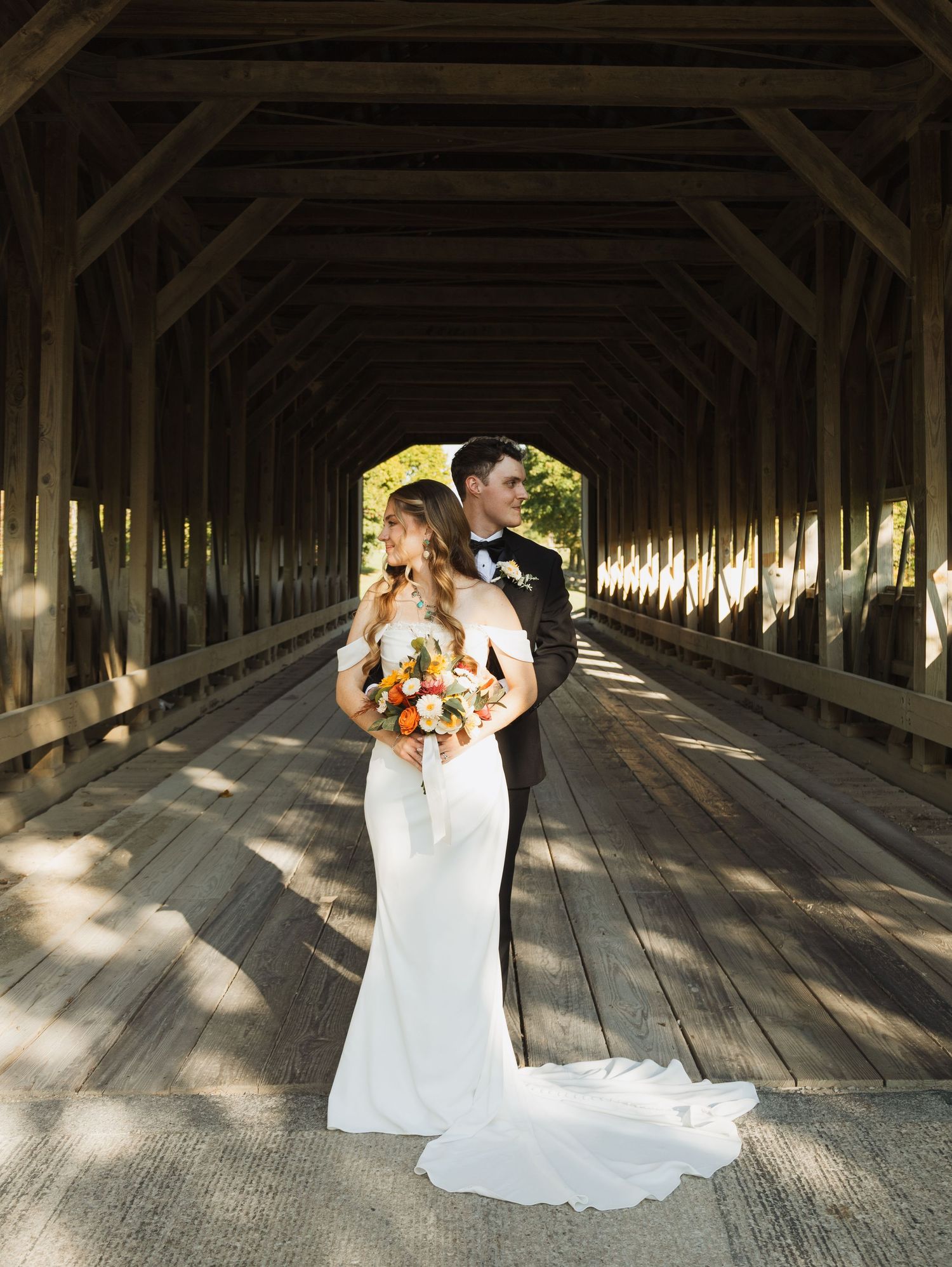 Wedding portrait inside a rustic covered wooden bridge with dramatic lighting and shadows.