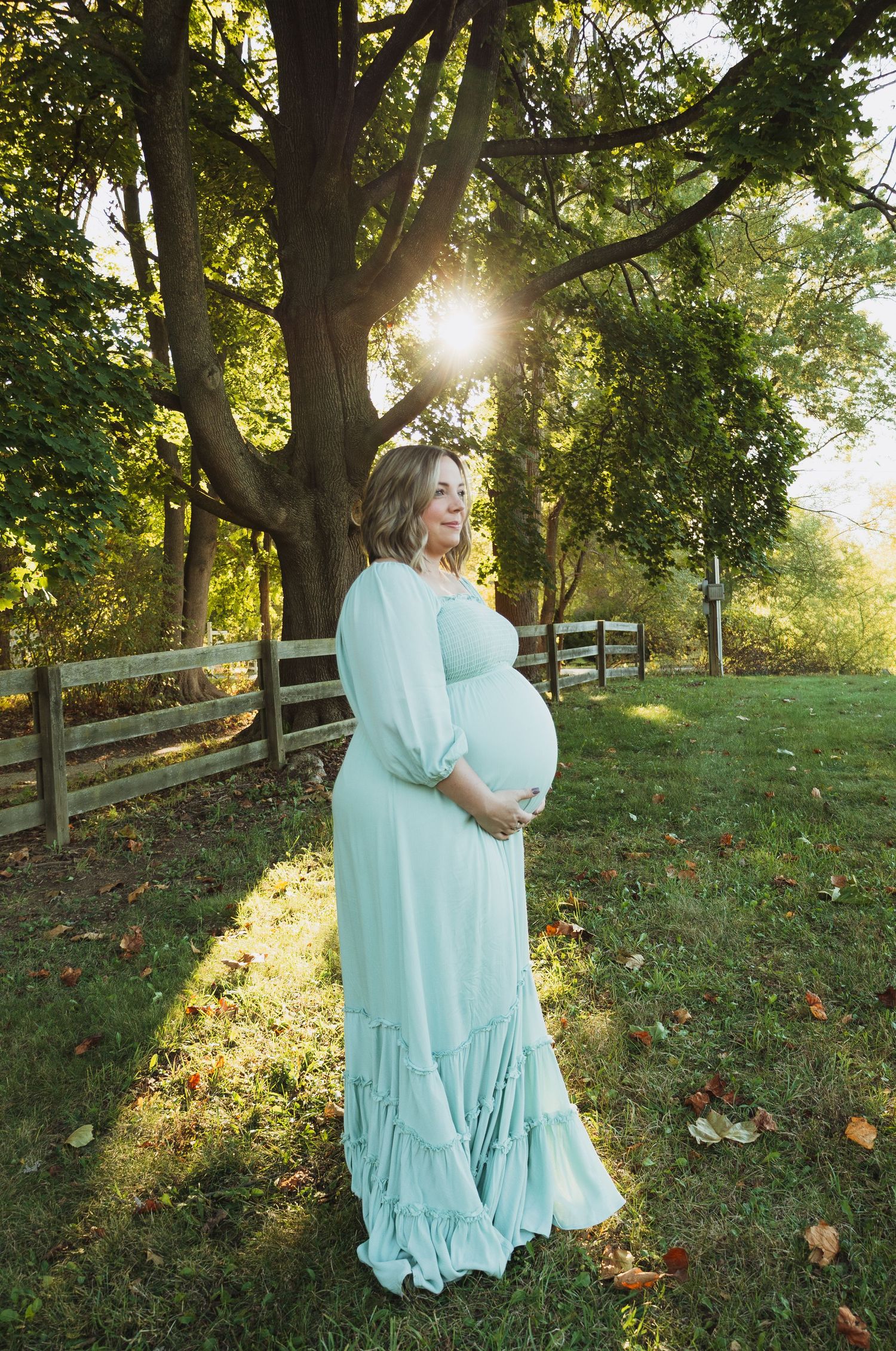 A pregnant woman in a flowing mint green dress poses in a sunlit park setting during golden hour.