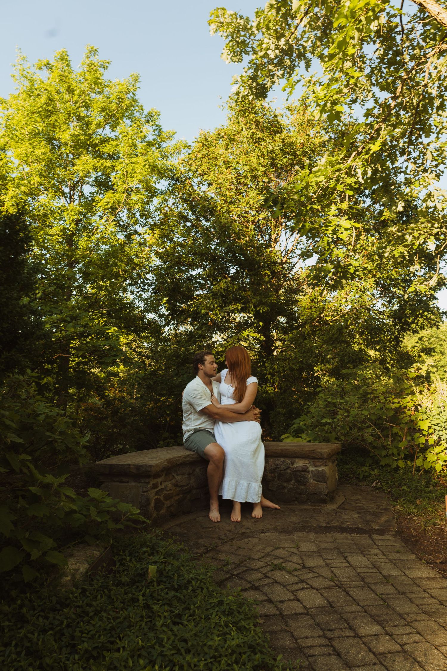 A couple shares a romantic kiss while sitting on a stone wall surrounded by lush green trees in a sunlit park.