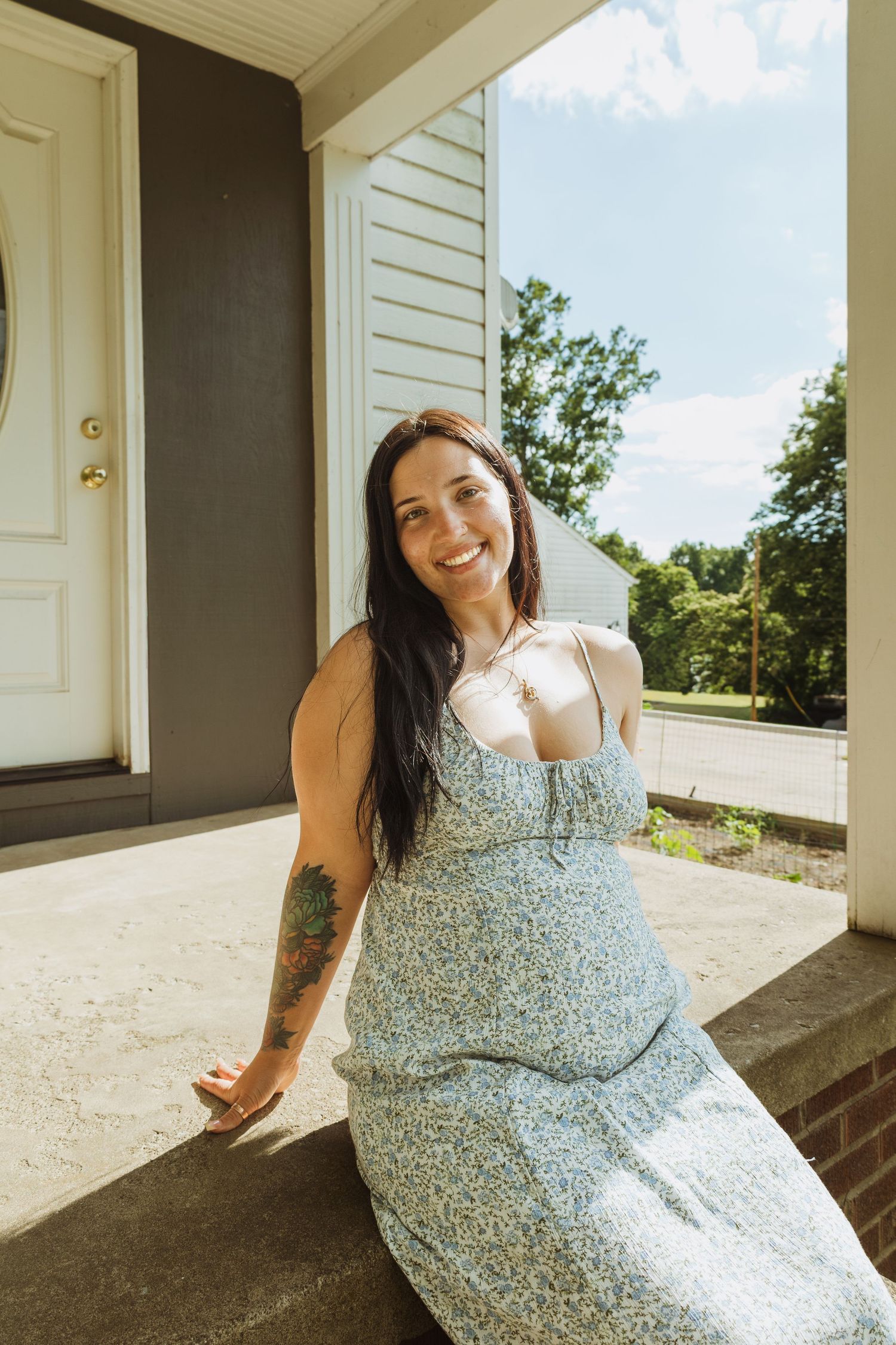 Smiling woman with dark hair and tattoos wearing a floral sundress, sitting on a sunny home porch.