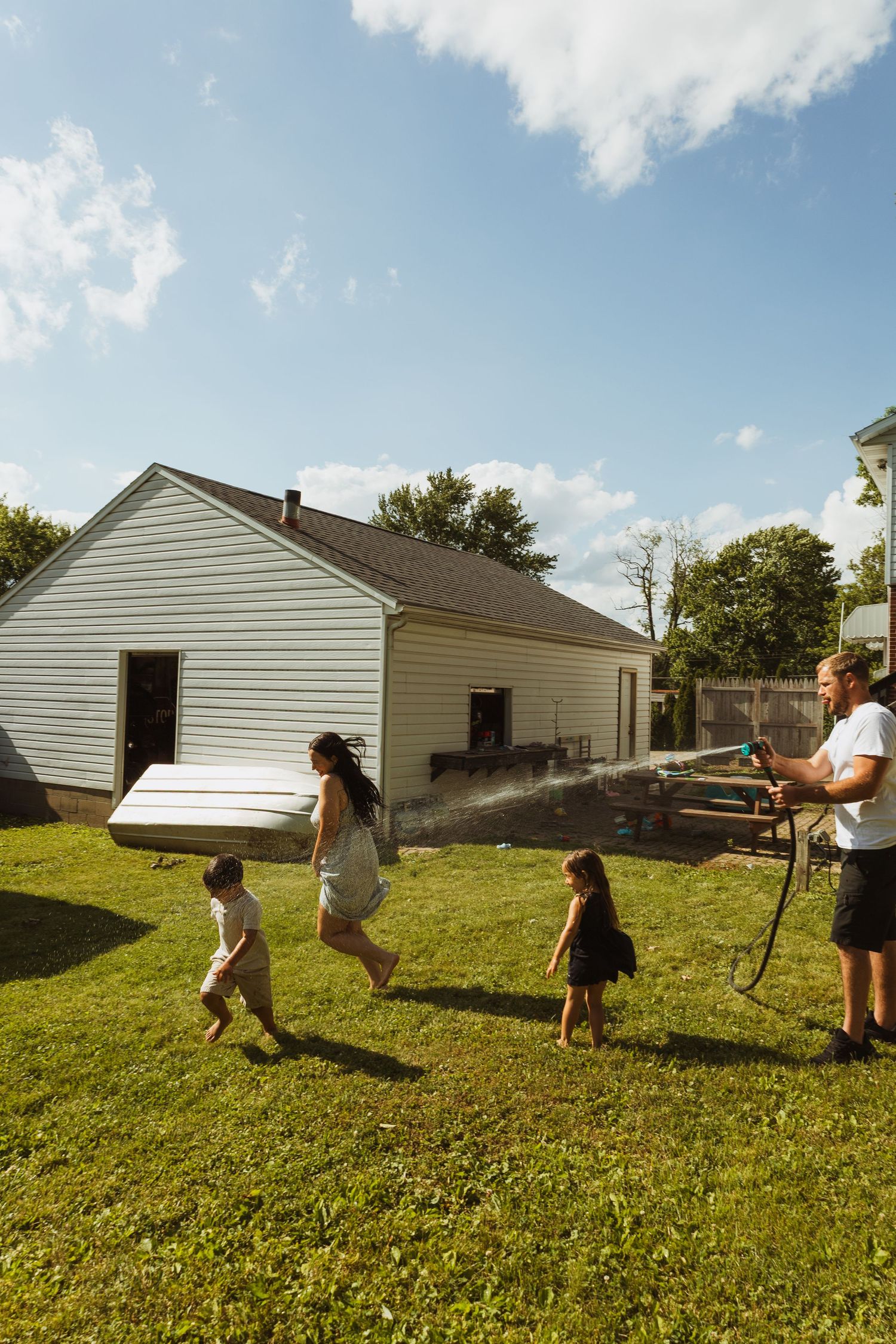 Family playing in backyard with garden hose on sunny day, children running on green grass near white house.
