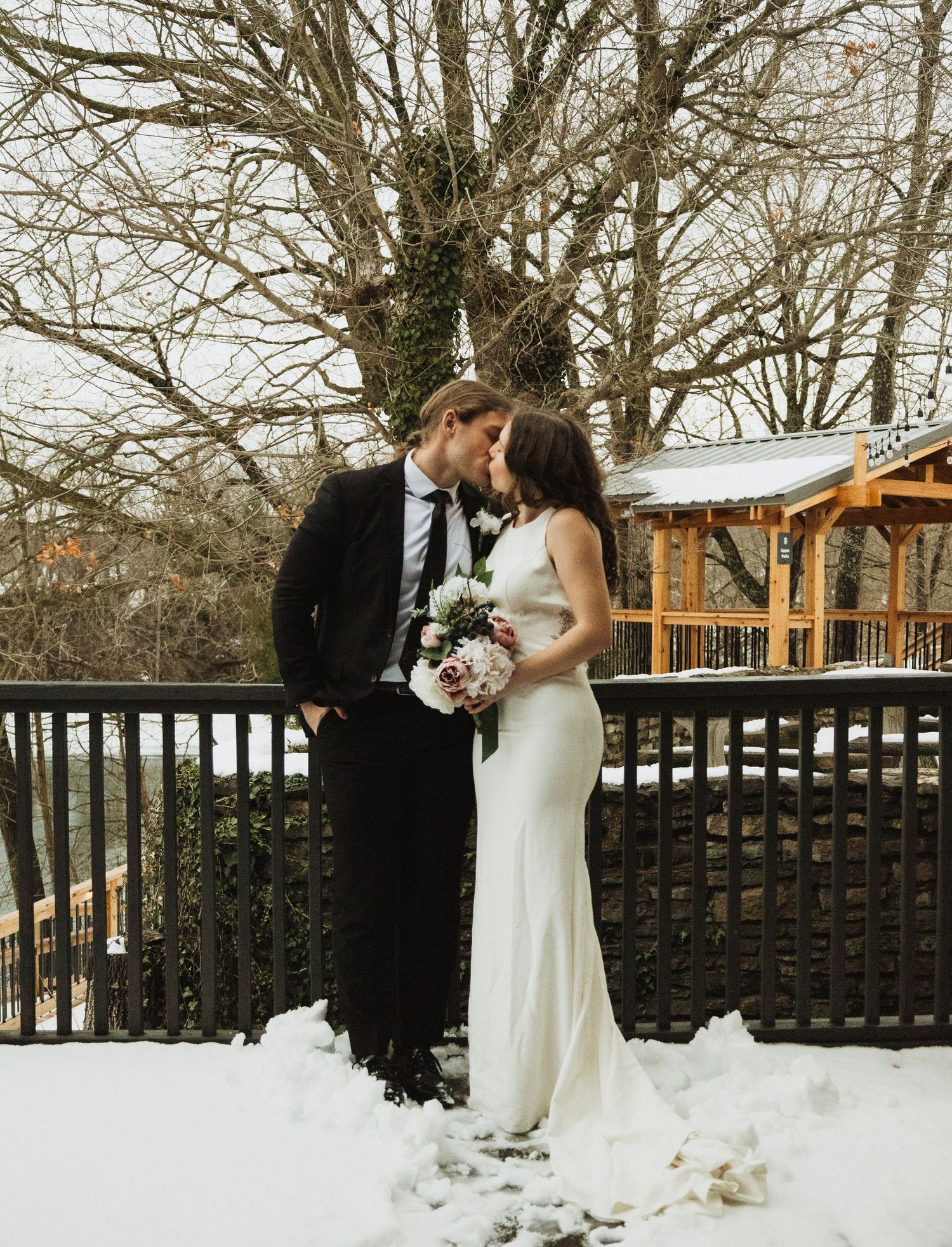 A winter wedding couple shares a romantic moment on a snow-covered deck with a rustic gazebo in the background.