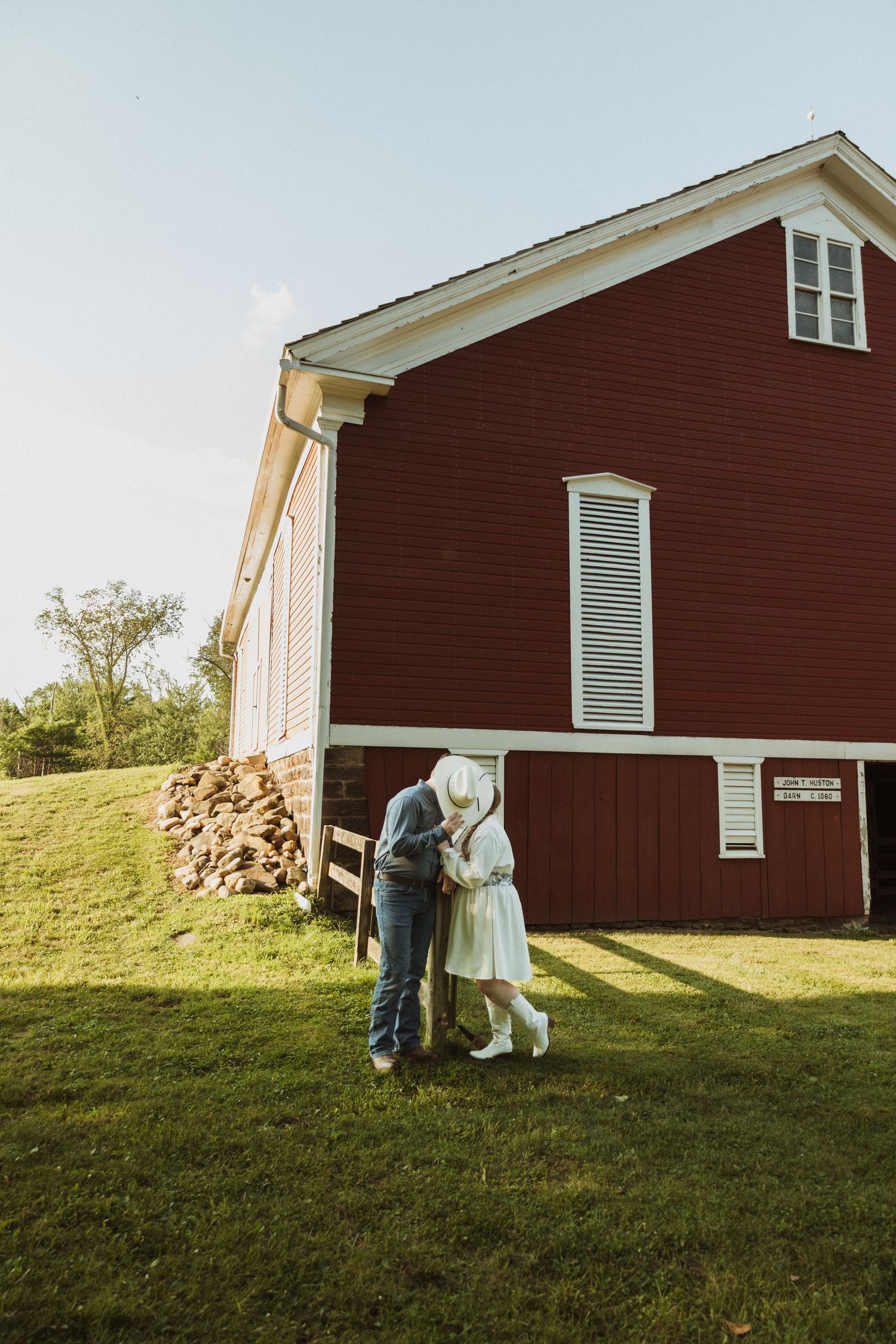 Two figures playfully interact near a rustic red barn on a grassy hillside in warm sunlight.