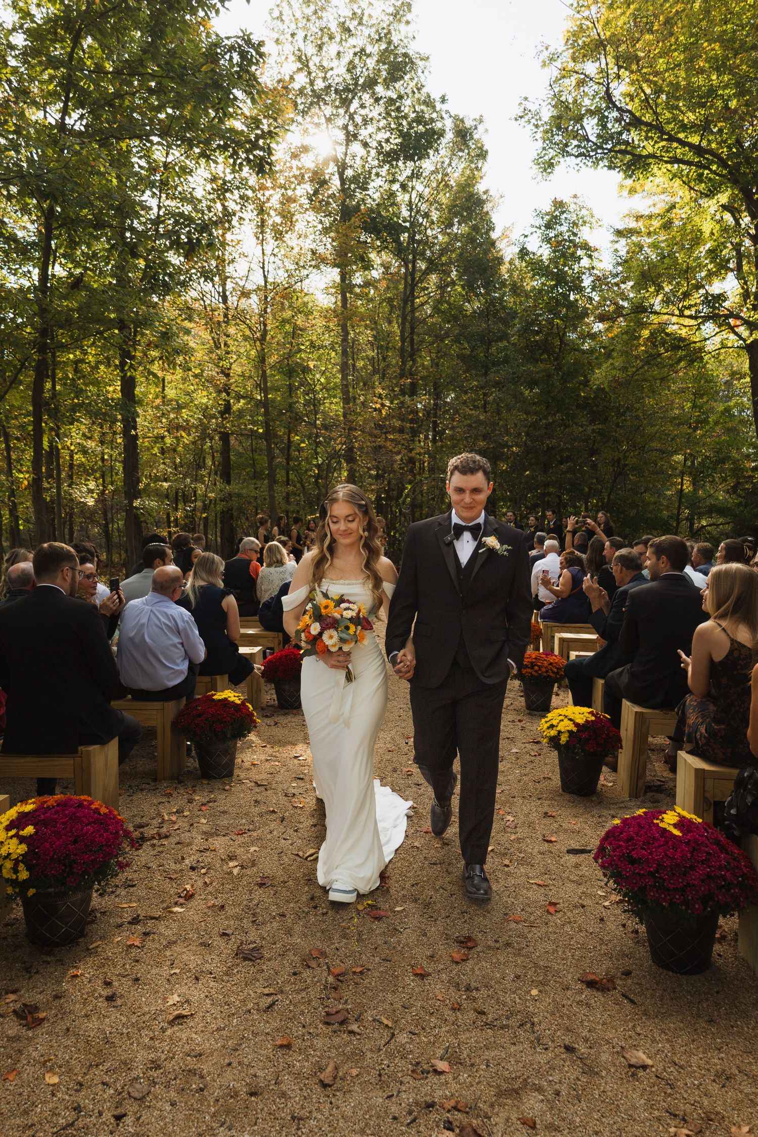 A wedding ceremony procession in a wooded outdoor setting with fall foliage and rustic wooden seating.