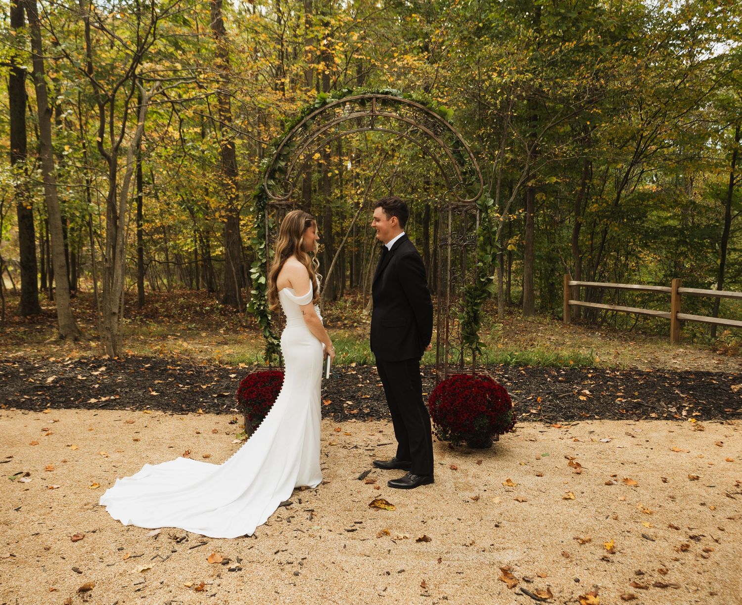 Bride and groom share first look moment under rustic arch surrounded by autumn trees.
