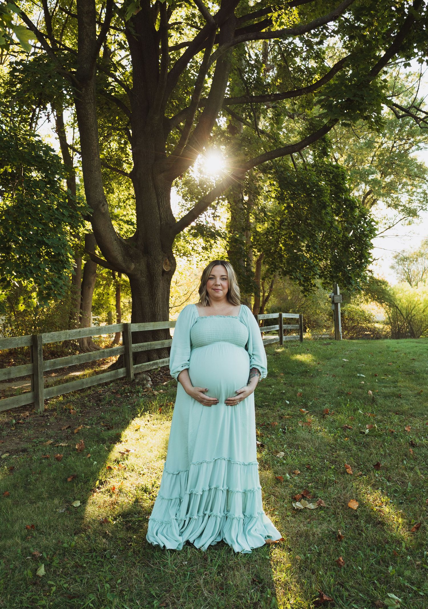 A serene maternity photo in a mint green maxi dress against a sunlit backdrop of trees and wooden fencing.