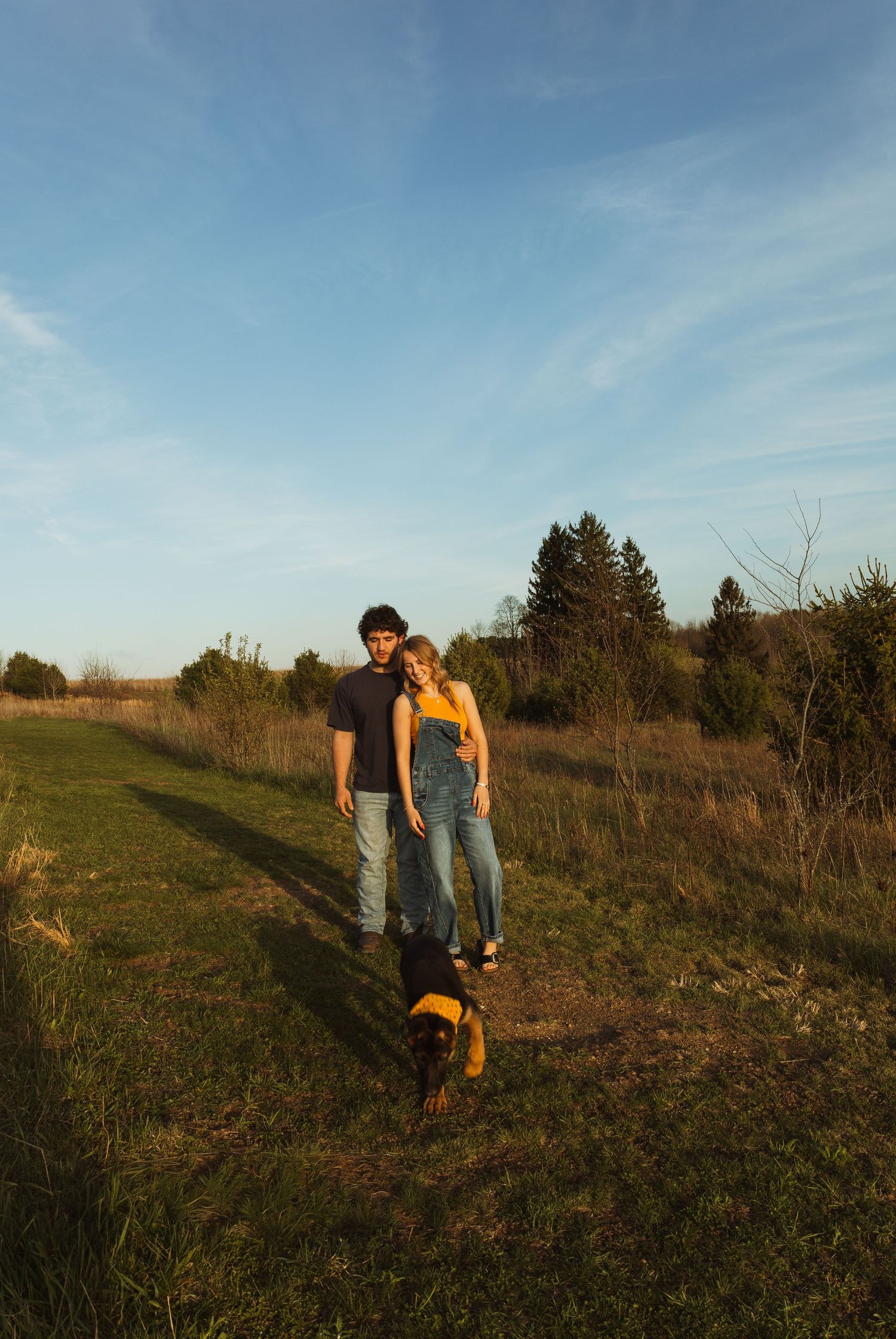 Couple walking with a Rottweiler dog on a grassy rural path at golden hour with trees and blue sky.