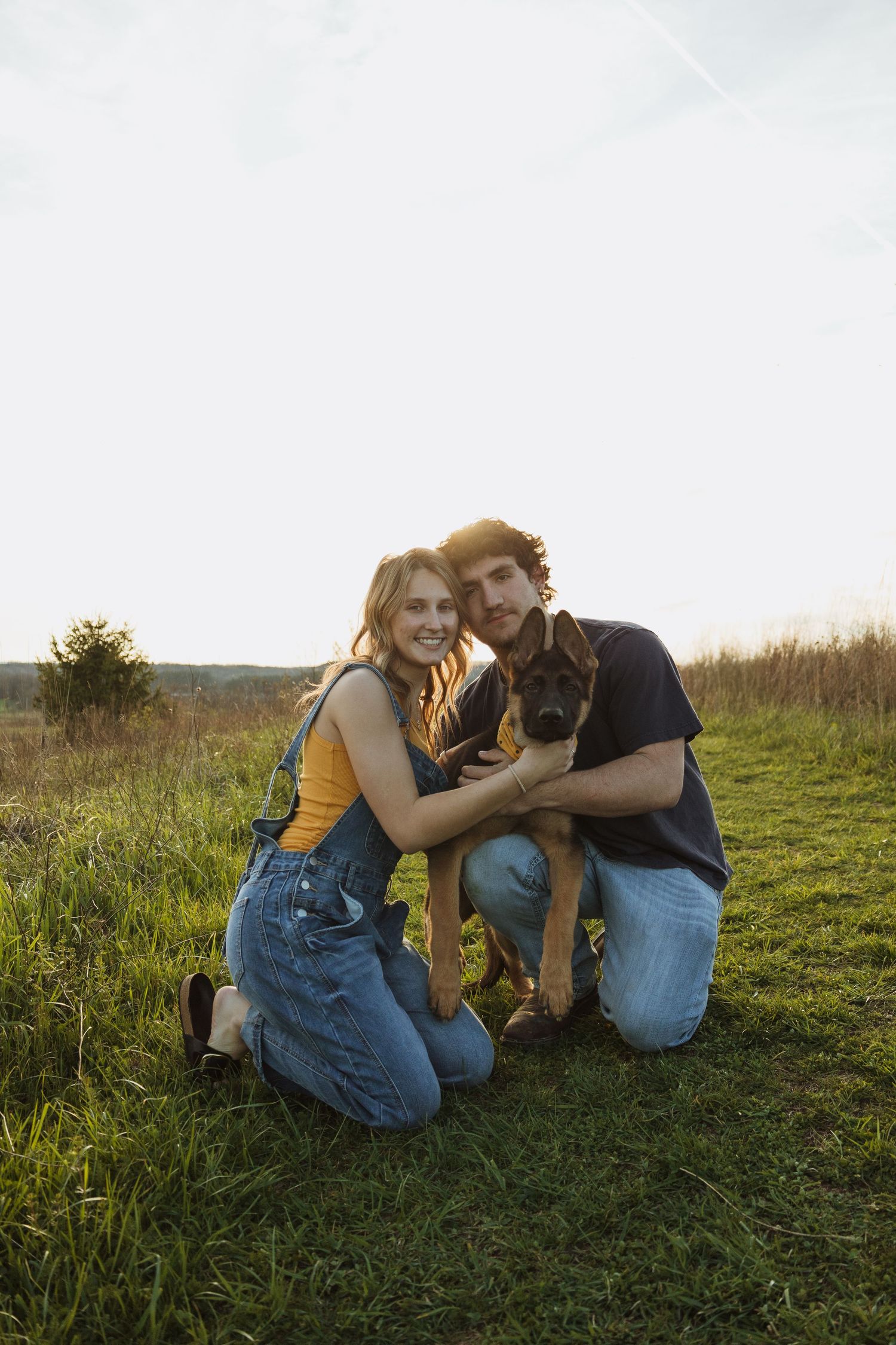 A smiling couple kneels on grass at sunset, holding an adorable puppy in a scenic field.