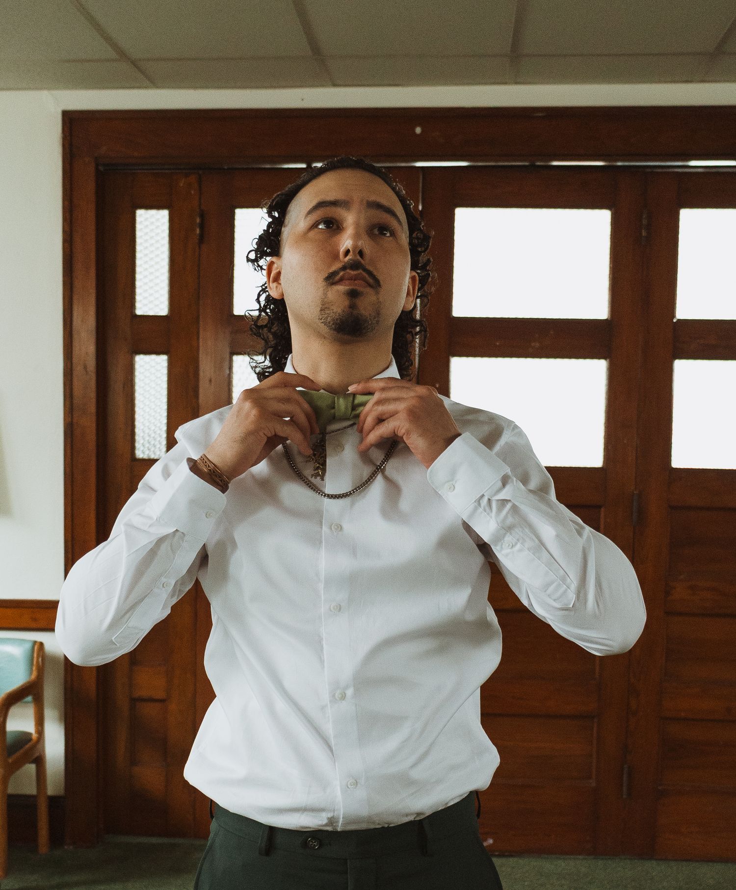 Man in white shirt adjusting green bow tie, with curly hair, standing in a room with wooden doors.