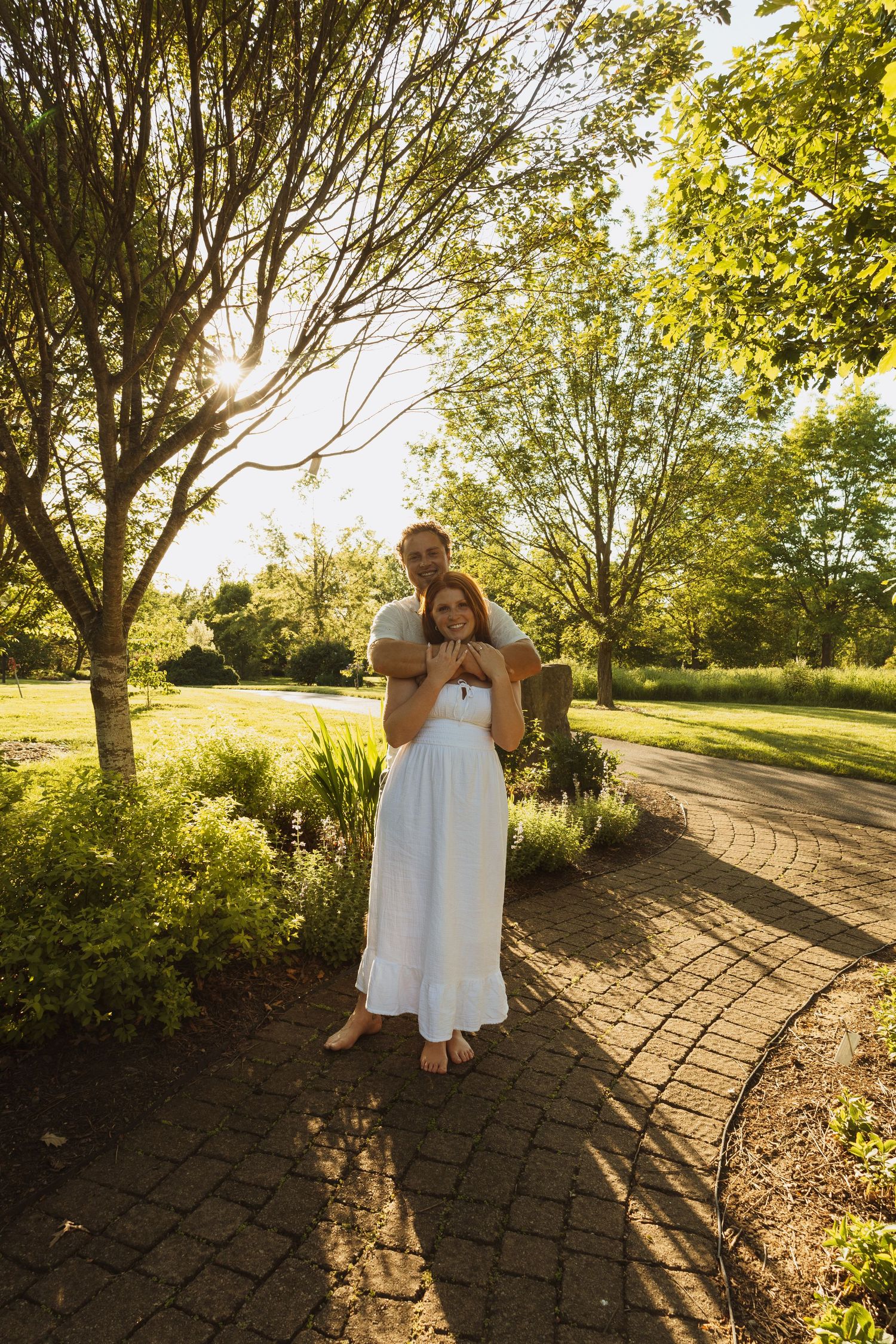 Couple embracing on a brick path in a sunny park, woman in white dress, man in gray shirt, surrounded by lush green trees.