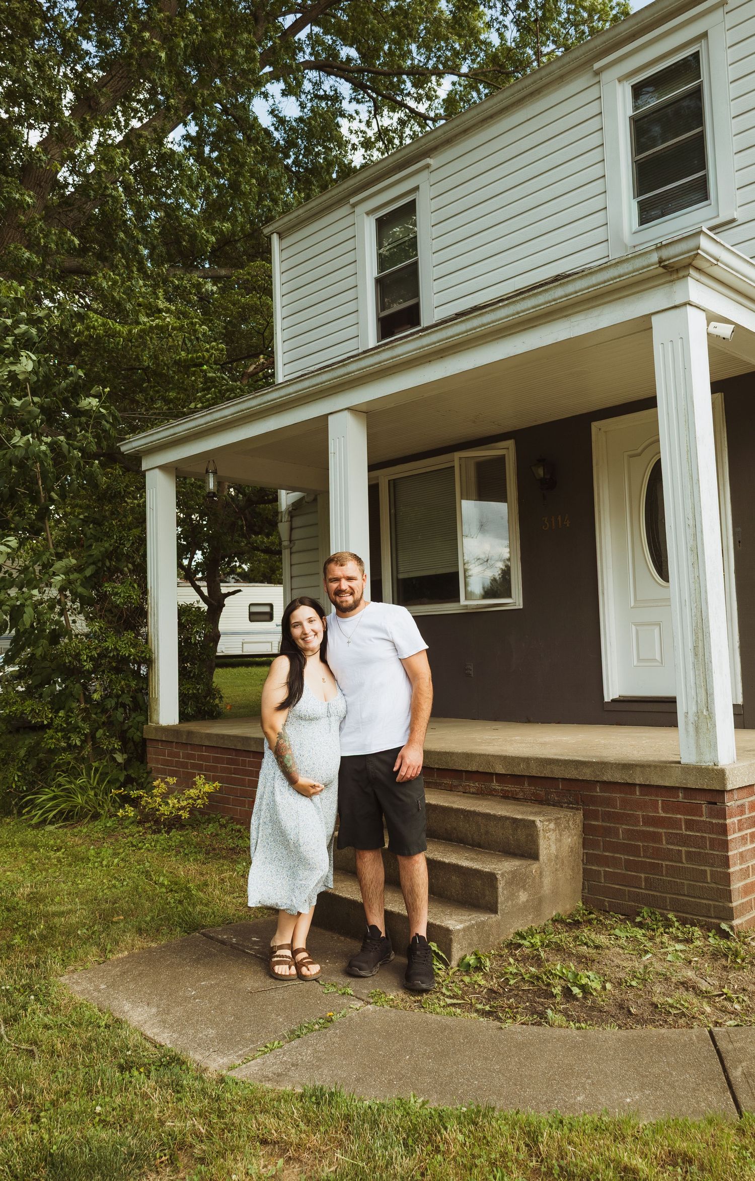 A smiling couple stands in front of a charming two-story home with a covered porch on a sunny day.
