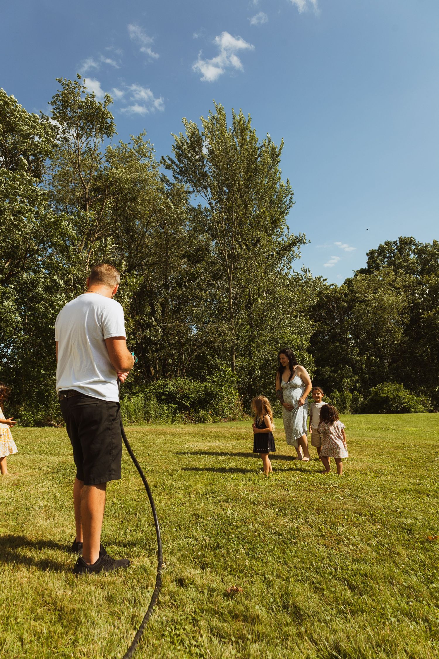 Family enjoying outdoor fun with a garden hose on a sunny green lawn surrounded by trees.