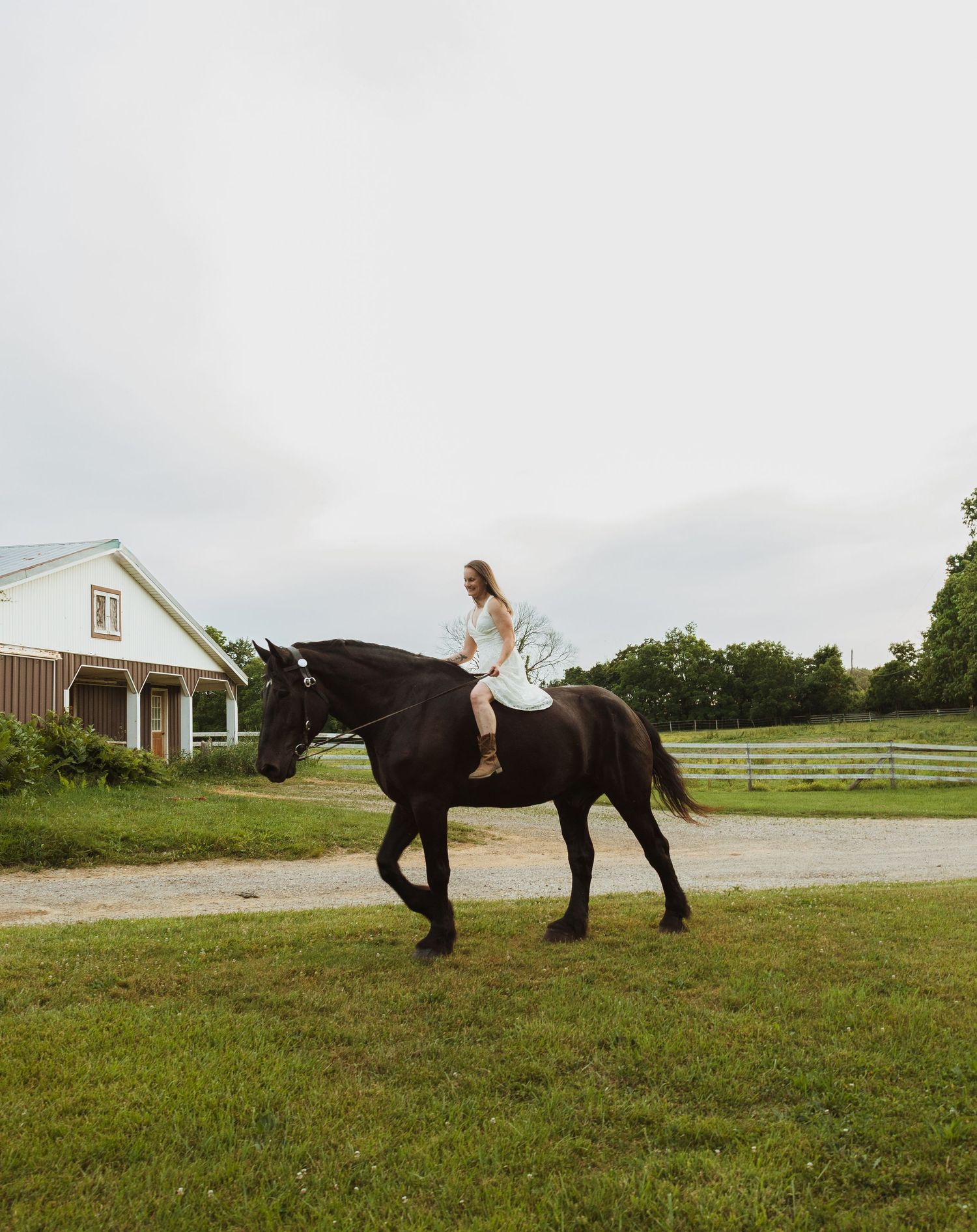 A woman in a white dress rides a black horse on a green farm with a white barn in the background.