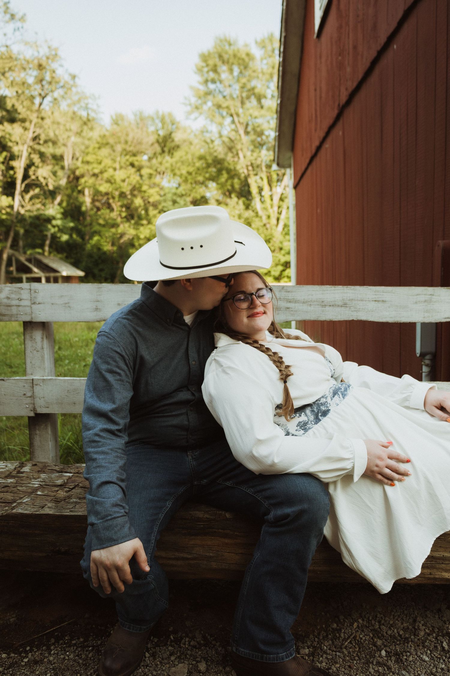 Couple in western attire share an intimate moment on a wooden bench at a rustic barn.