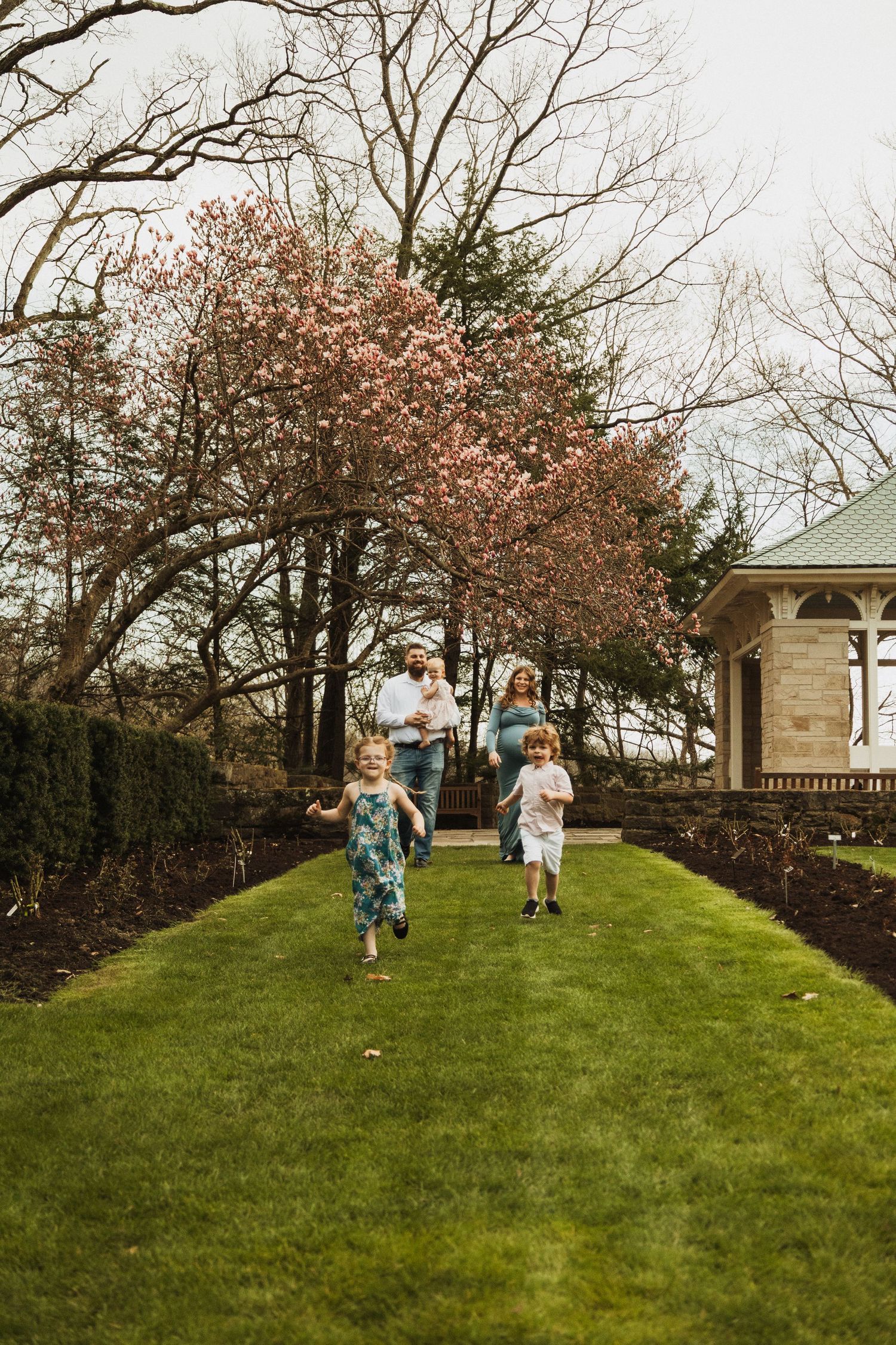 A group joyfully runs down a grassy path bordered by flowering trees and a gazebo on a spring day.