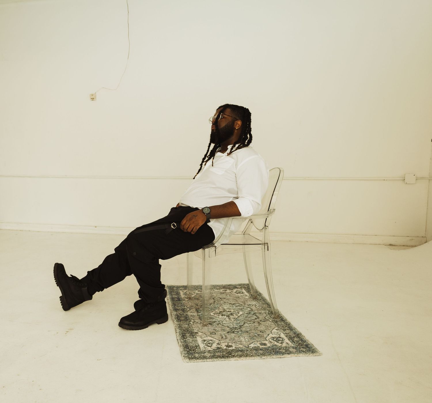 Person with dreadlocks sits sideways on a white chair over an ornate rug in an empty white room.