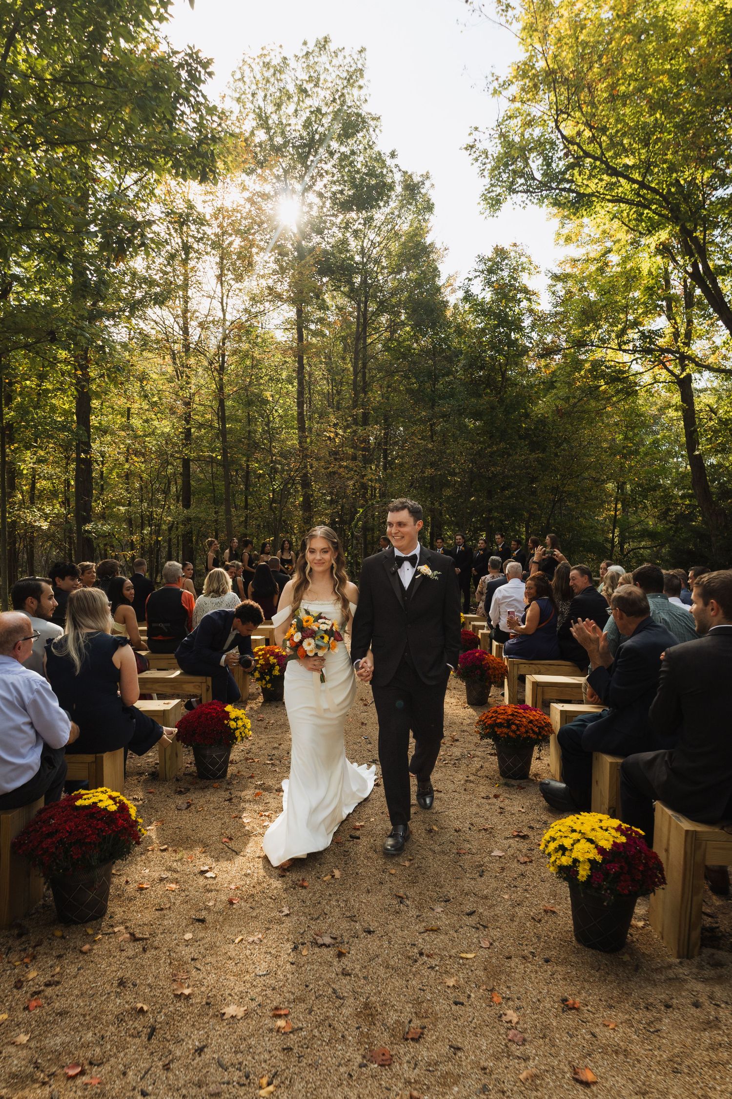 Autumn outdoor wedding ceremony with guests seated on wooden benches surrounded by fall foliage.