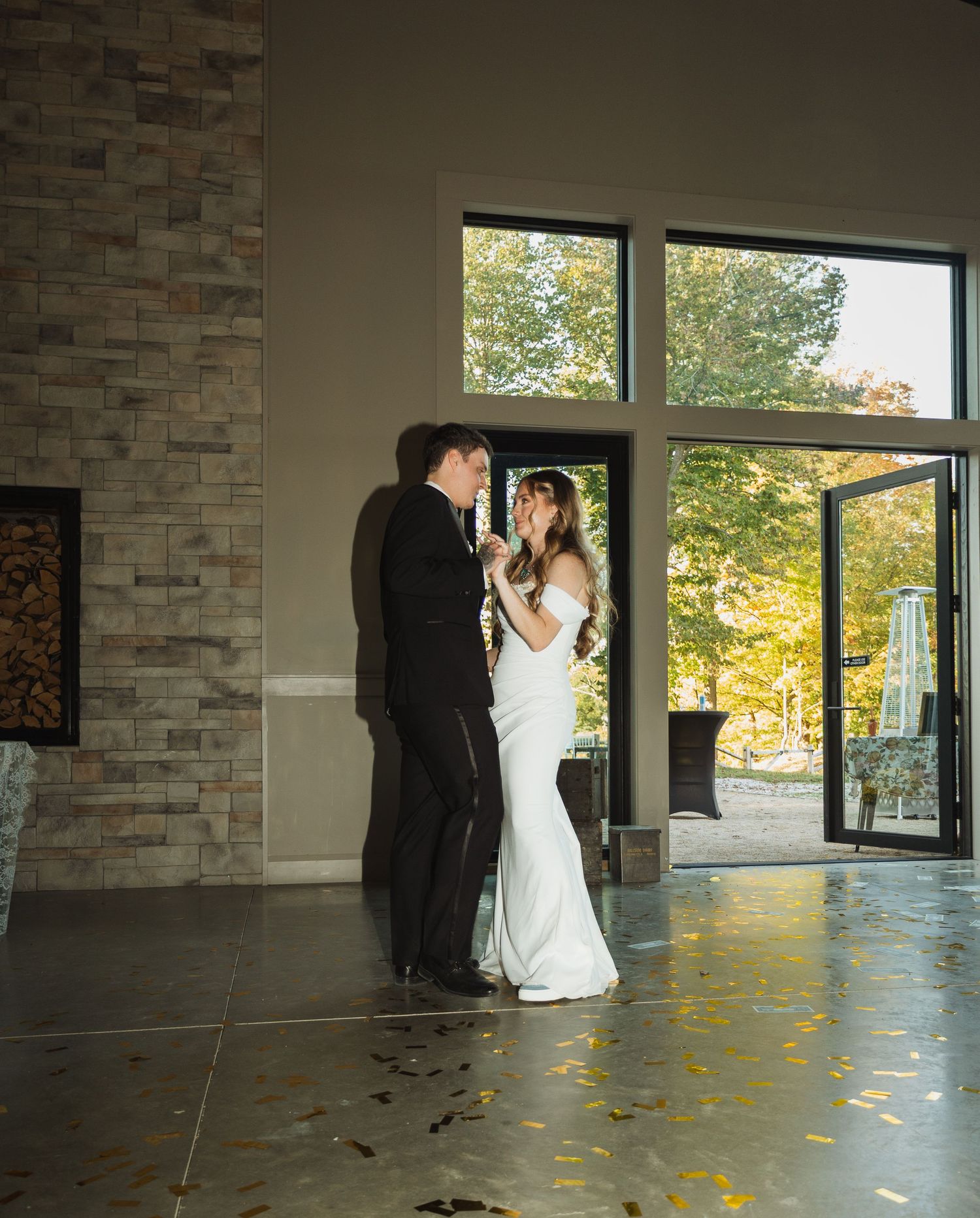 Couple dancing in sunlit room with stone walls and large windows overlooking autumn trees.
