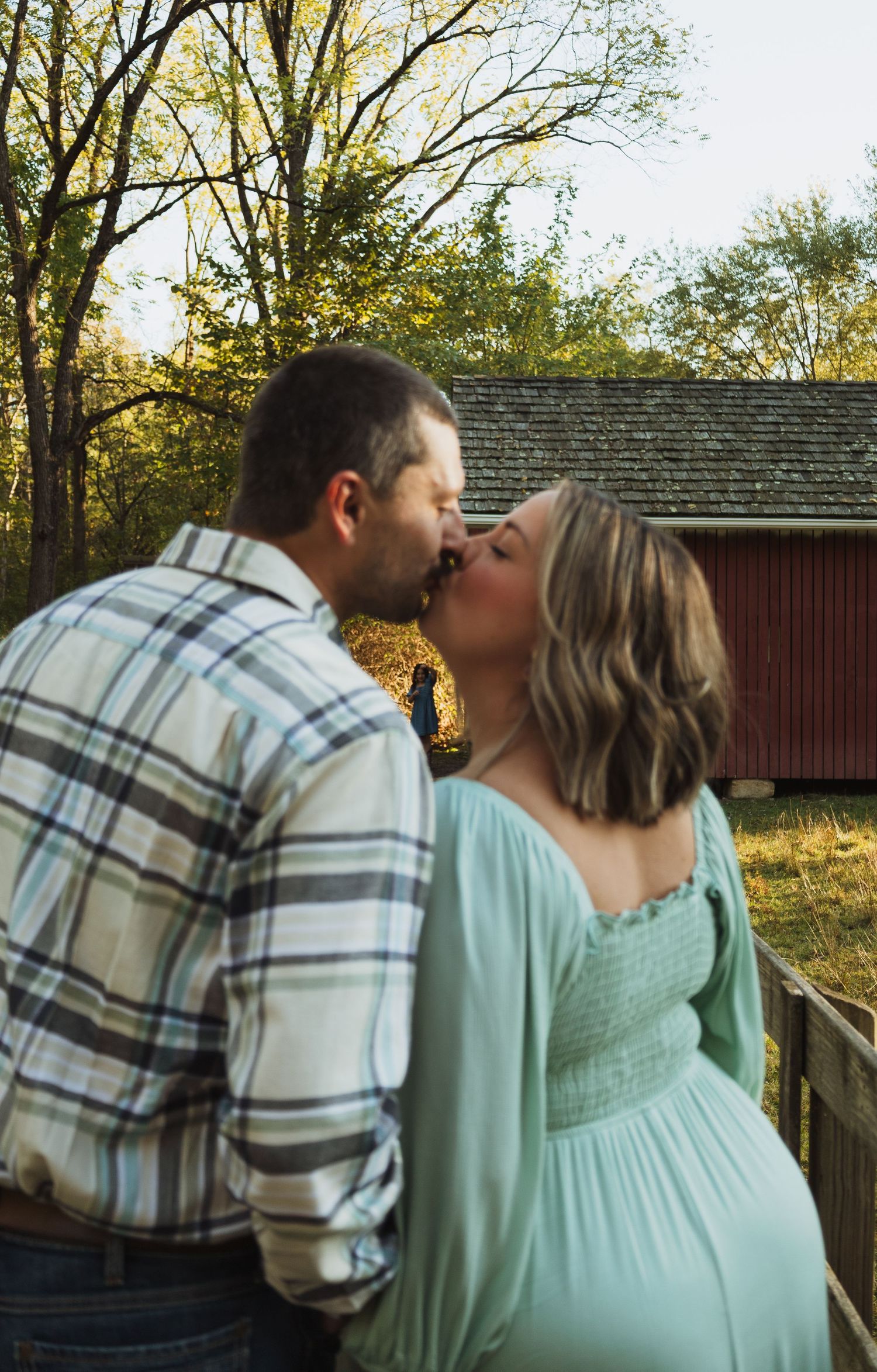 A couple in plaid shirt and mint green dress share a tender moment outdoors in autumn sunlight.