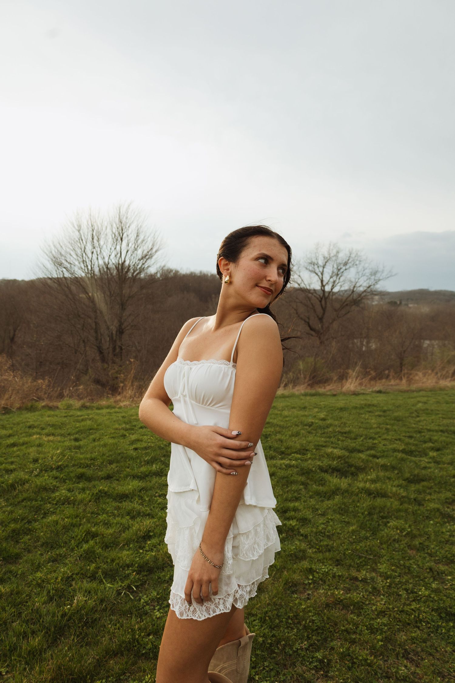 Young woman in white ruffled dress standing in green field with bare trees and overcast sky in background.