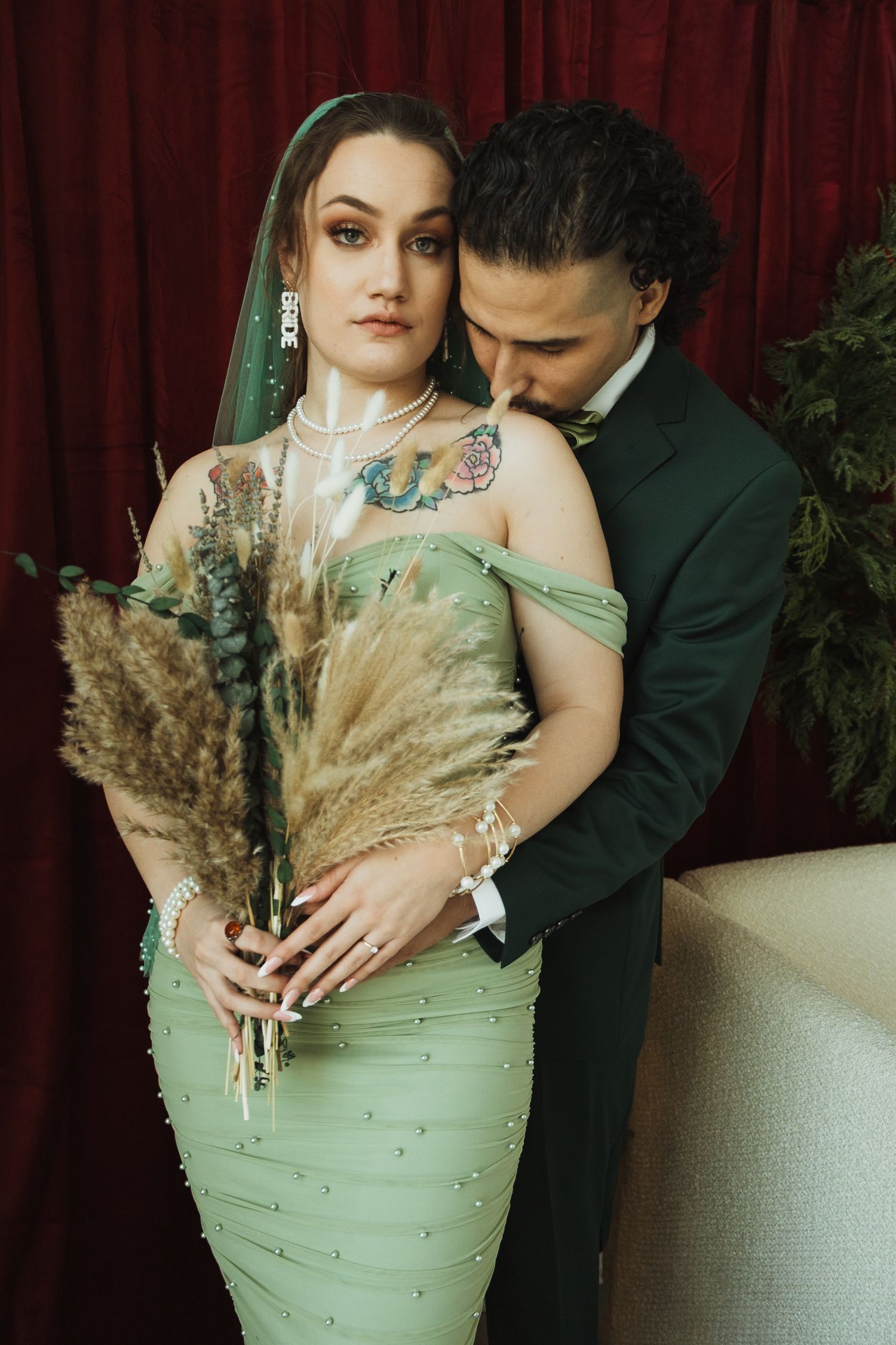 Groom in dark green suit embracing tattooed bride holding pampas grass bouquet against red drape.