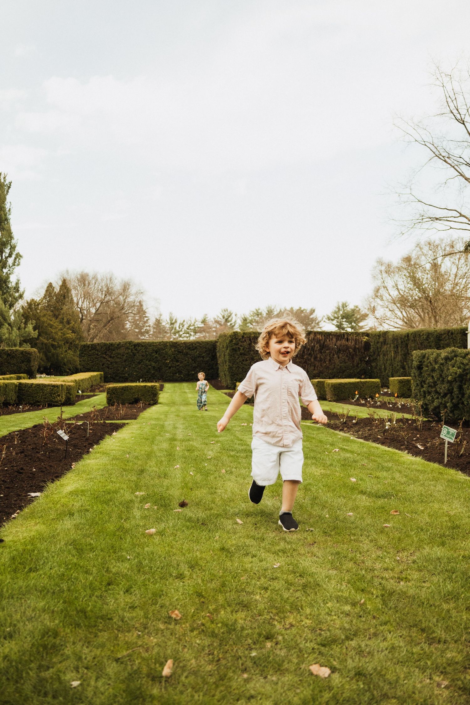 A child runs joyfully through a manicured garden with neatly trimmed hedges and well-maintained lawn paths.
