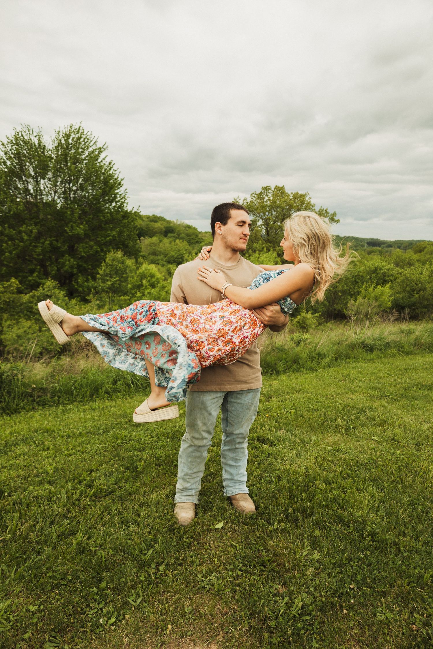 A romantic couple embraces in a scenic field with green rolling hills and overcast skies in the background.